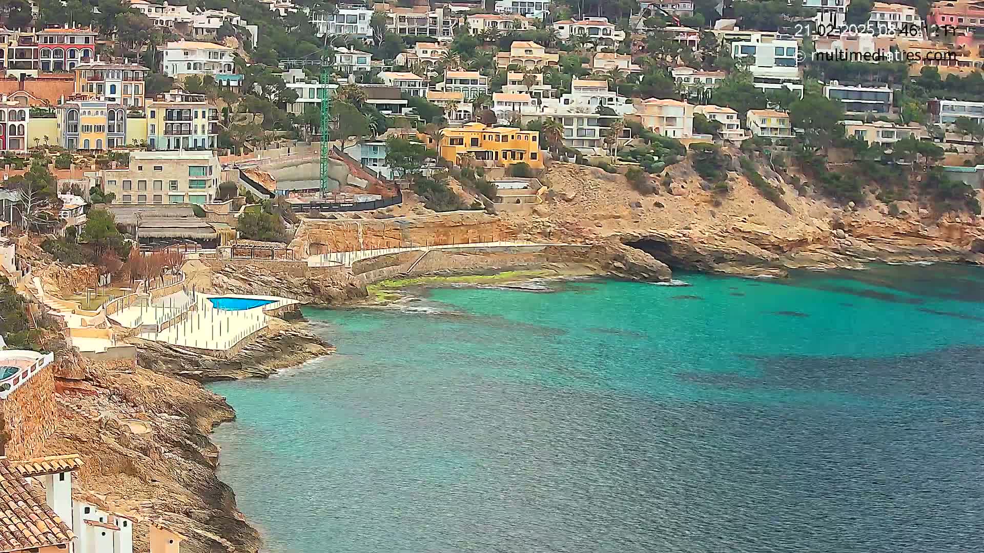 A coastal scene shows a collection of houses built on a hillside overlooking a calm, teal-colored sea, with a small pool and patio area near the water's edge.