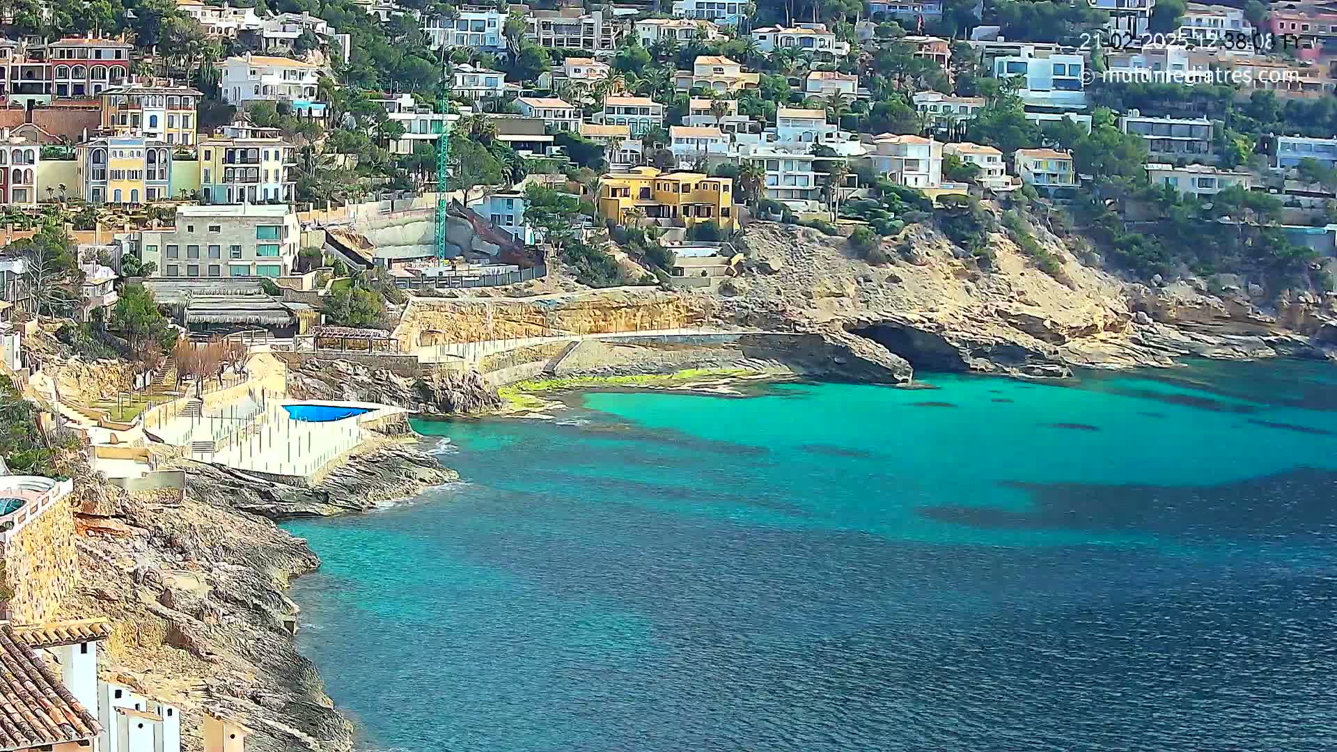 A coastal town with numerous houses clings to a rocky hillside overlooking a vibrant turquoise sea on a sunny day.
