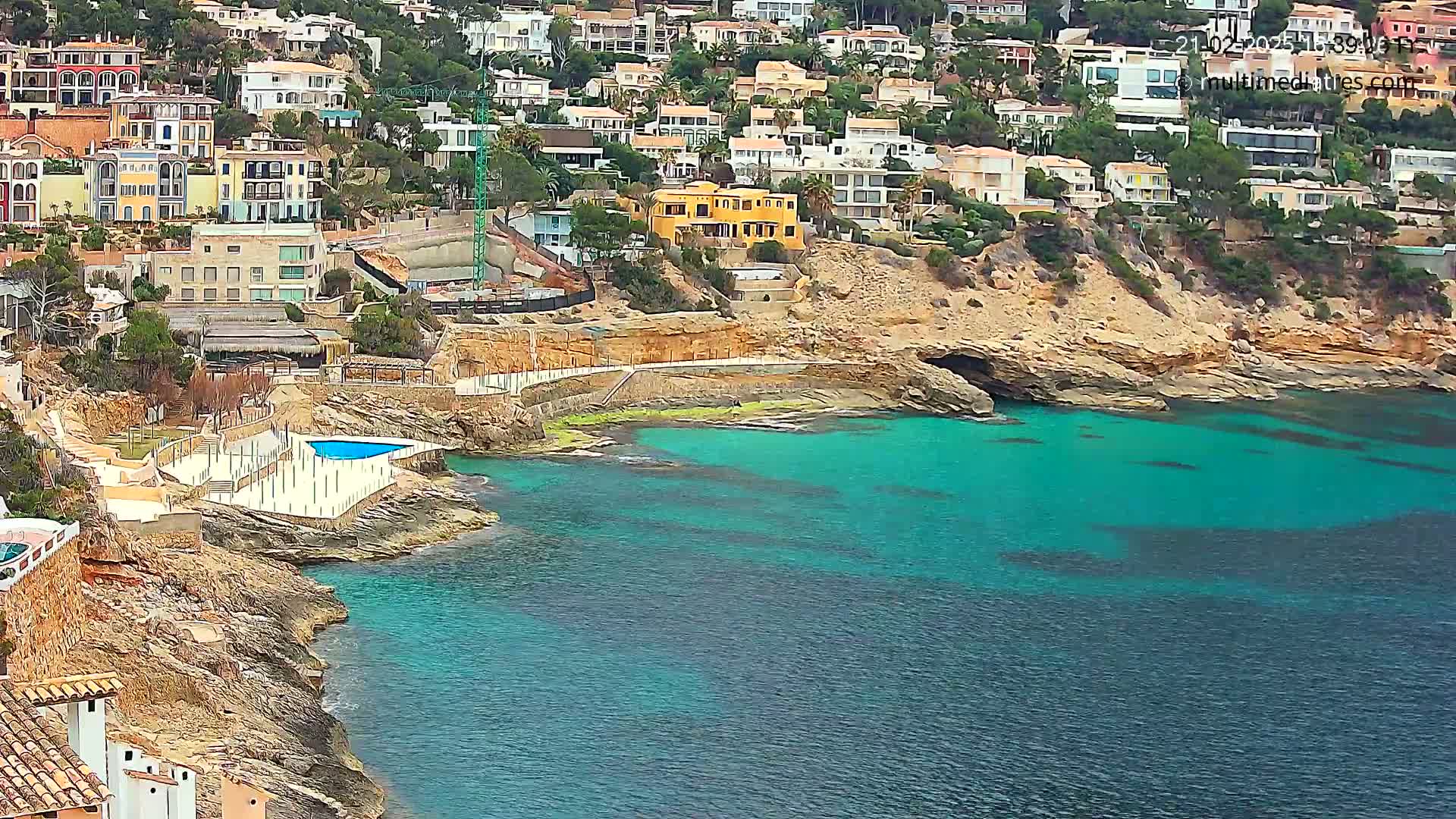 A rocky coastline with turquoise water features a small pool and many houses built into the hillside.