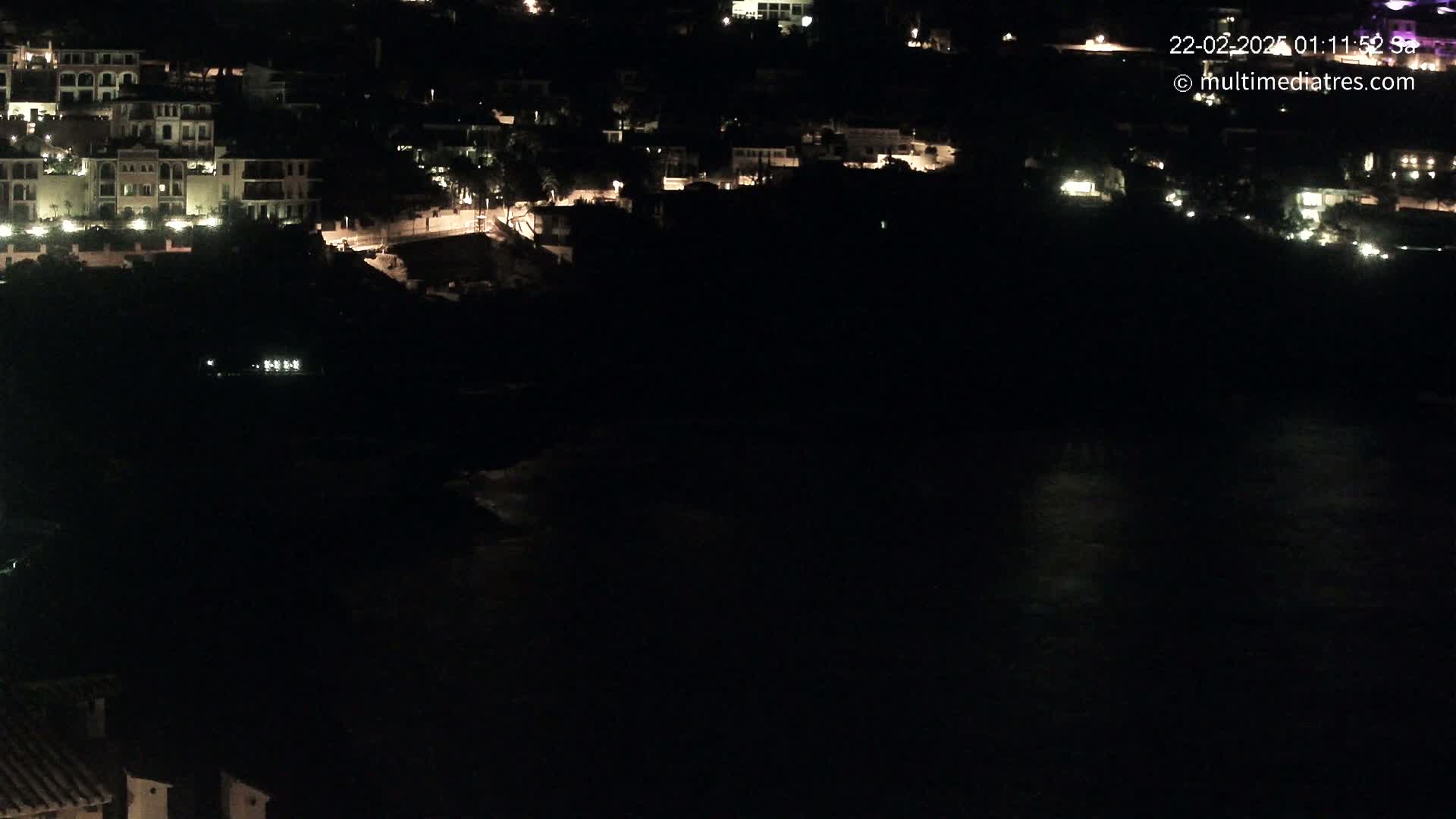 A nighttime, aerial view shows a dark body of water below a collection of dimly lit buildings on a hillside.