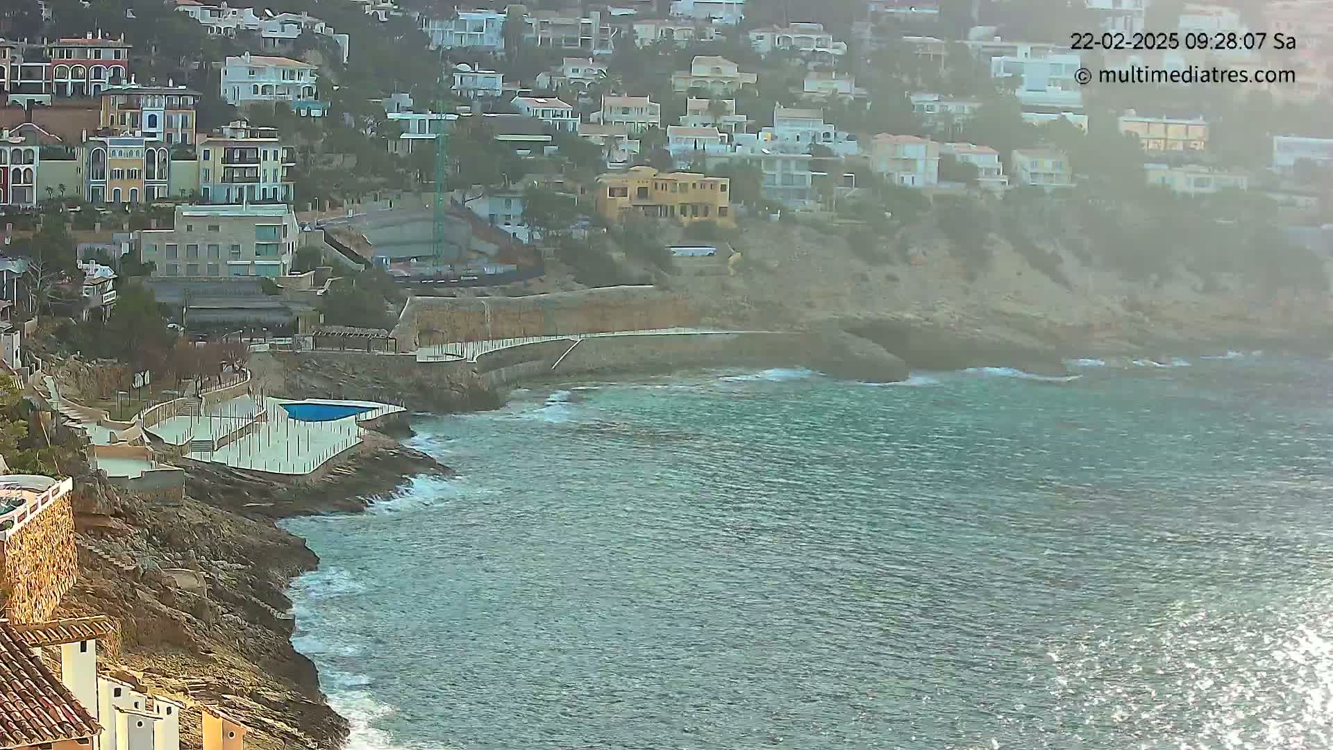 A coastal town built into a cliffside overlooks a calm, somewhat hazy sea with small waves lapping against the rocky shore, featuring a small pool near the water's edge.