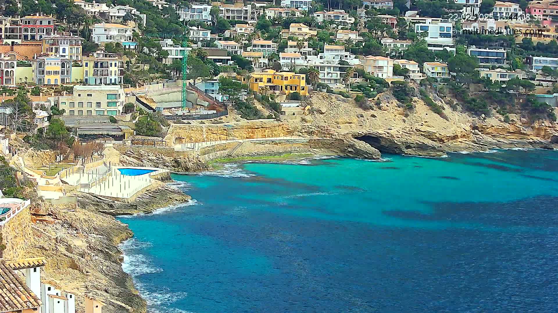 A rocky coastline with colorful houses perched on a hillside overlooks a vibrant turquoise sea with small waves.