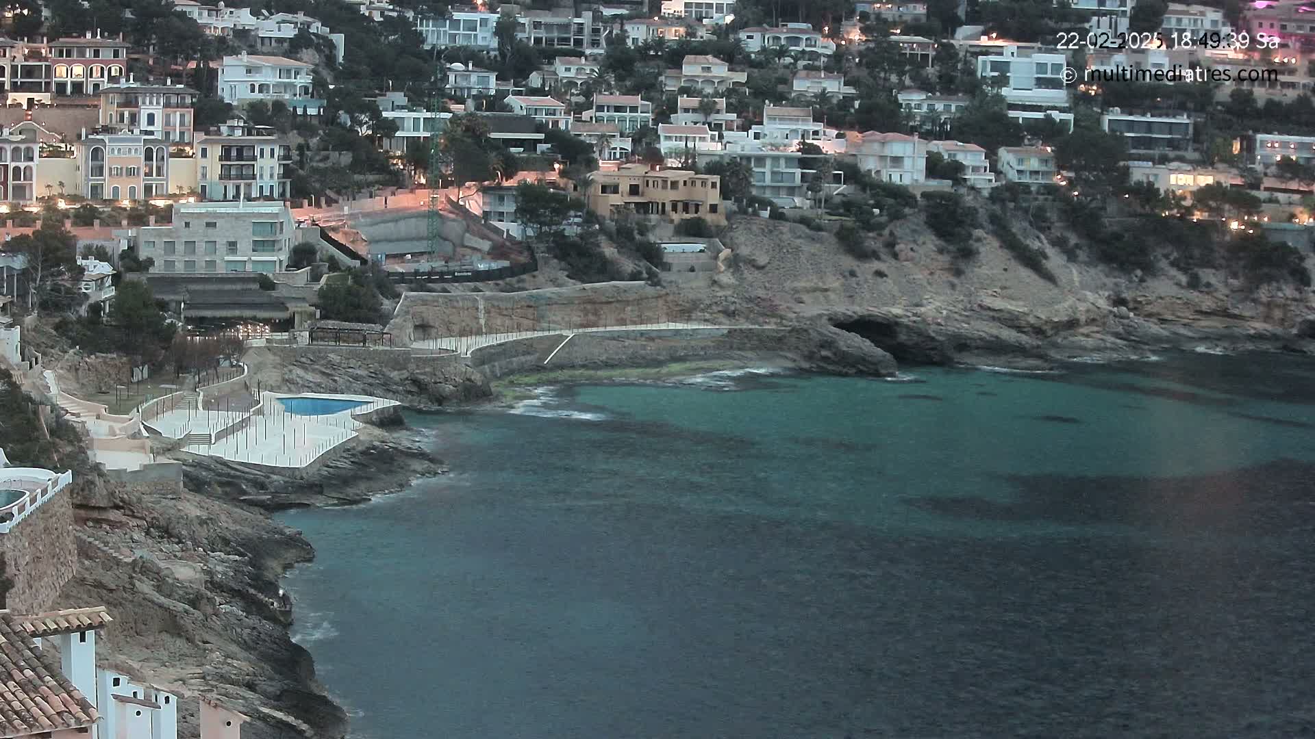 A rocky coastline with a small, calm, teal-colored bay features a collection of white buildings nestled on a hillside at twilight.