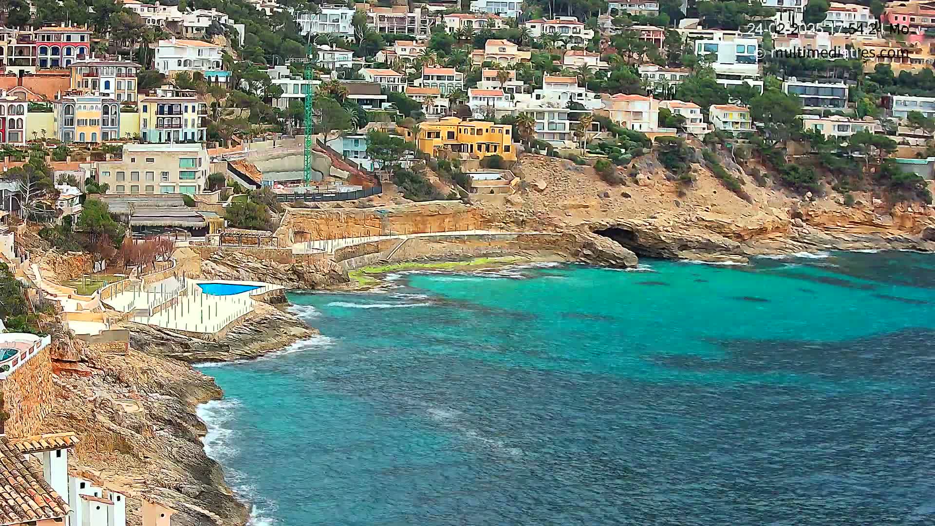 A rocky coastline with turquoise water features numerous houses built on a hillside, and a small swimming pool is visible near the water's edge.