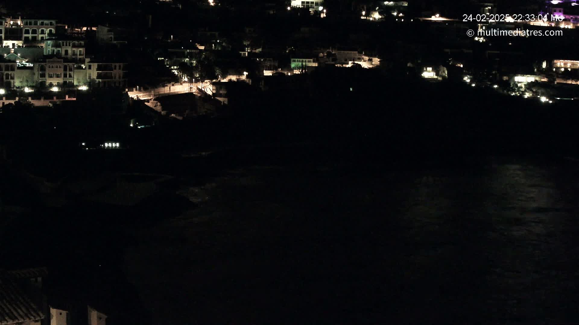 A nighttime, coastal view shows a collection of buildings with scattered lights overlooking a dark body of water.