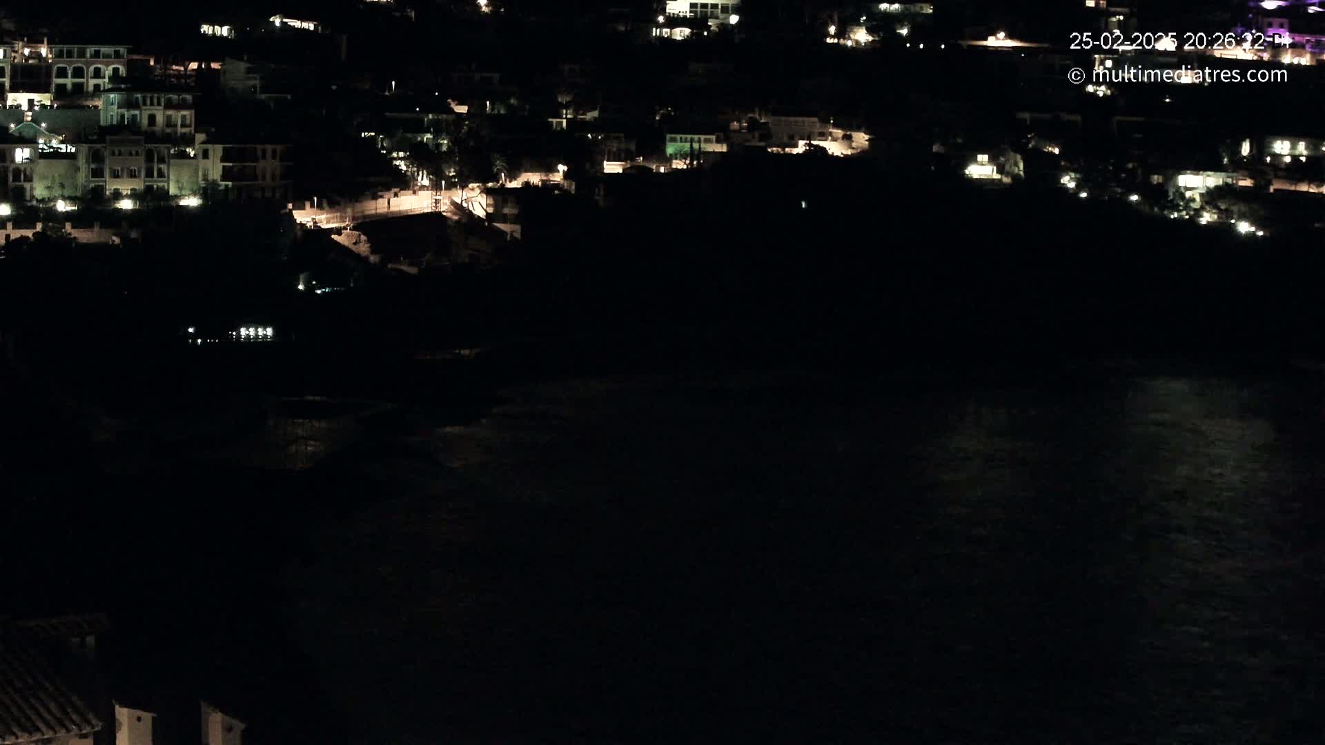 A nighttime coastal scene shows a dark body of water reflecting faint lights from a hillside village.