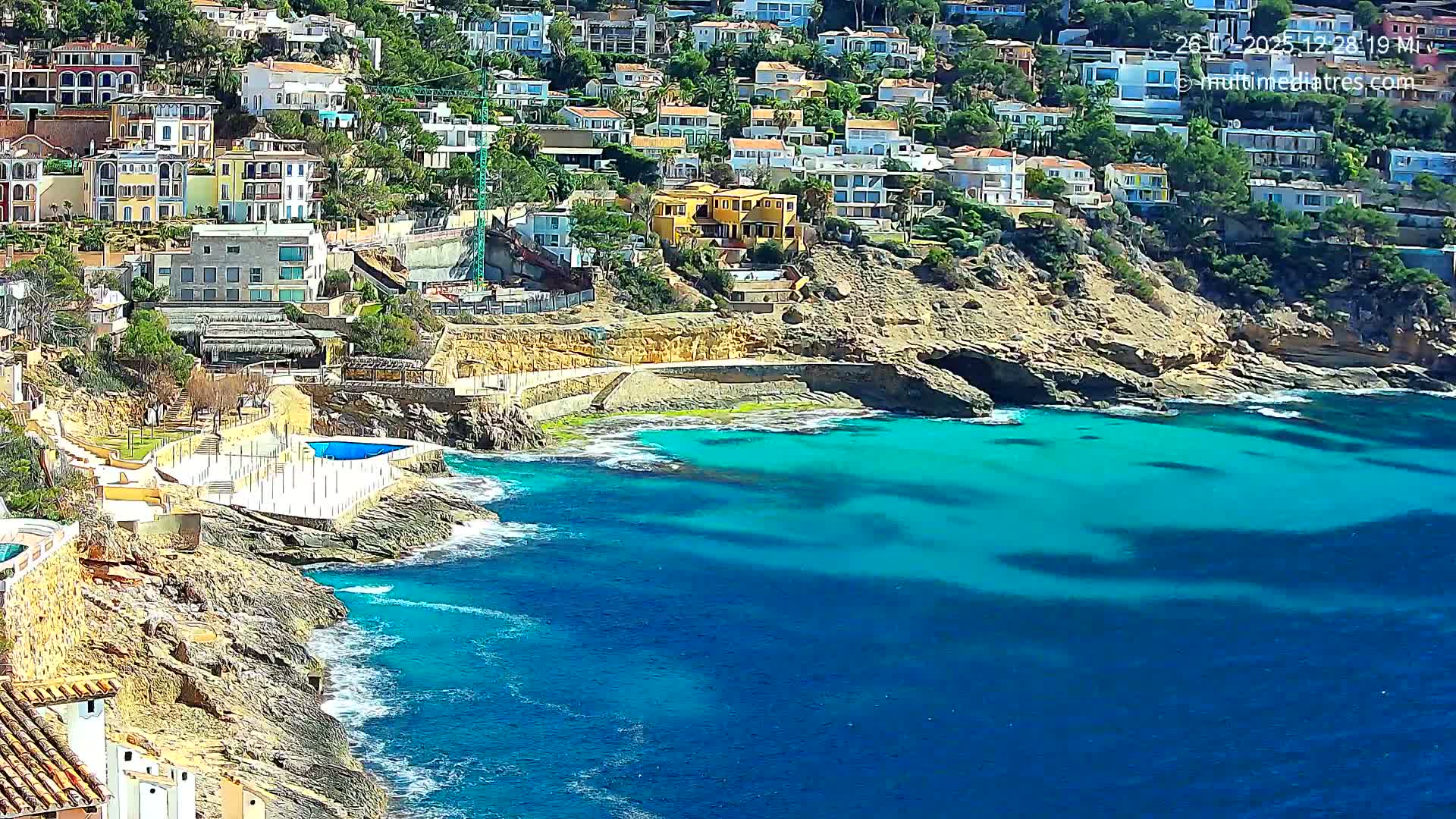 A hillside of colorful houses overlooks a rocky cove with turquoise water and small waves on a sunny day.