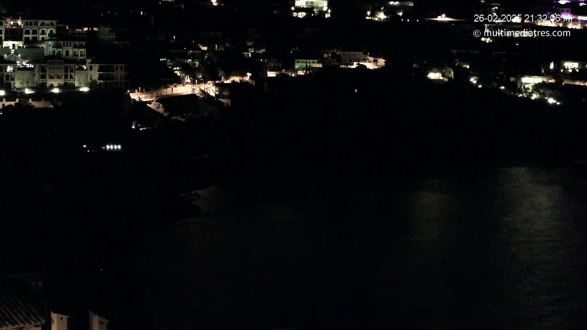 A nighttime view of a coastal town with dimly lit buildings and a dark body of water reflecting some light.