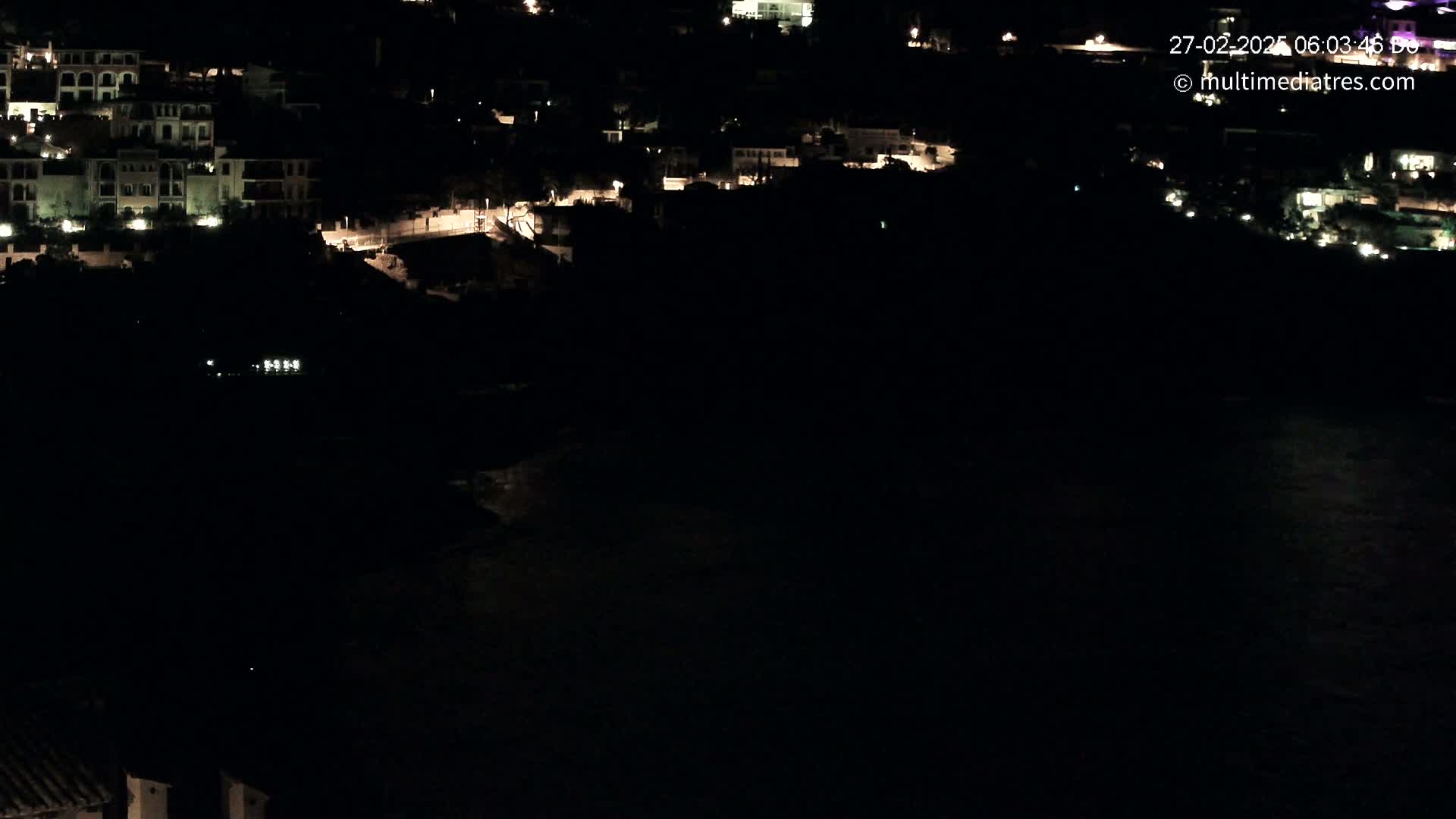 A nighttime long shot shows a coastal town with dimly lit buildings and a dark body of water below.