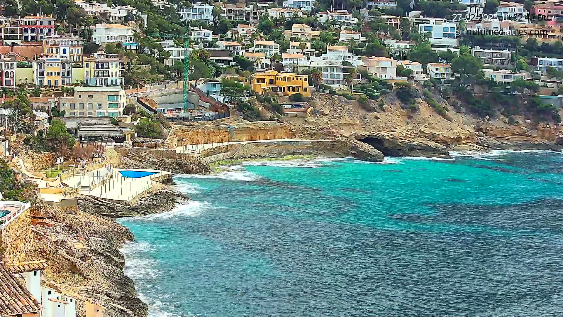 A rocky coastline with turquoise water features numerous houses built into a hillside, a small pool, and some light wave action.