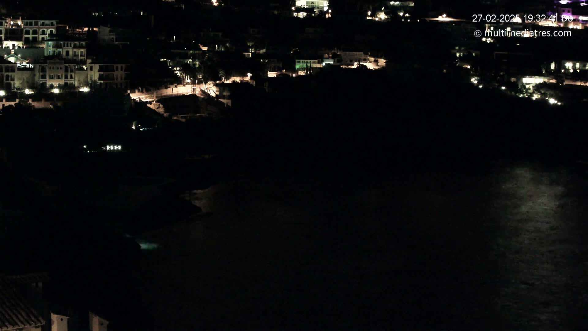 A nighttime view of a coastal town with lights illuminating buildings and a dark body of water.