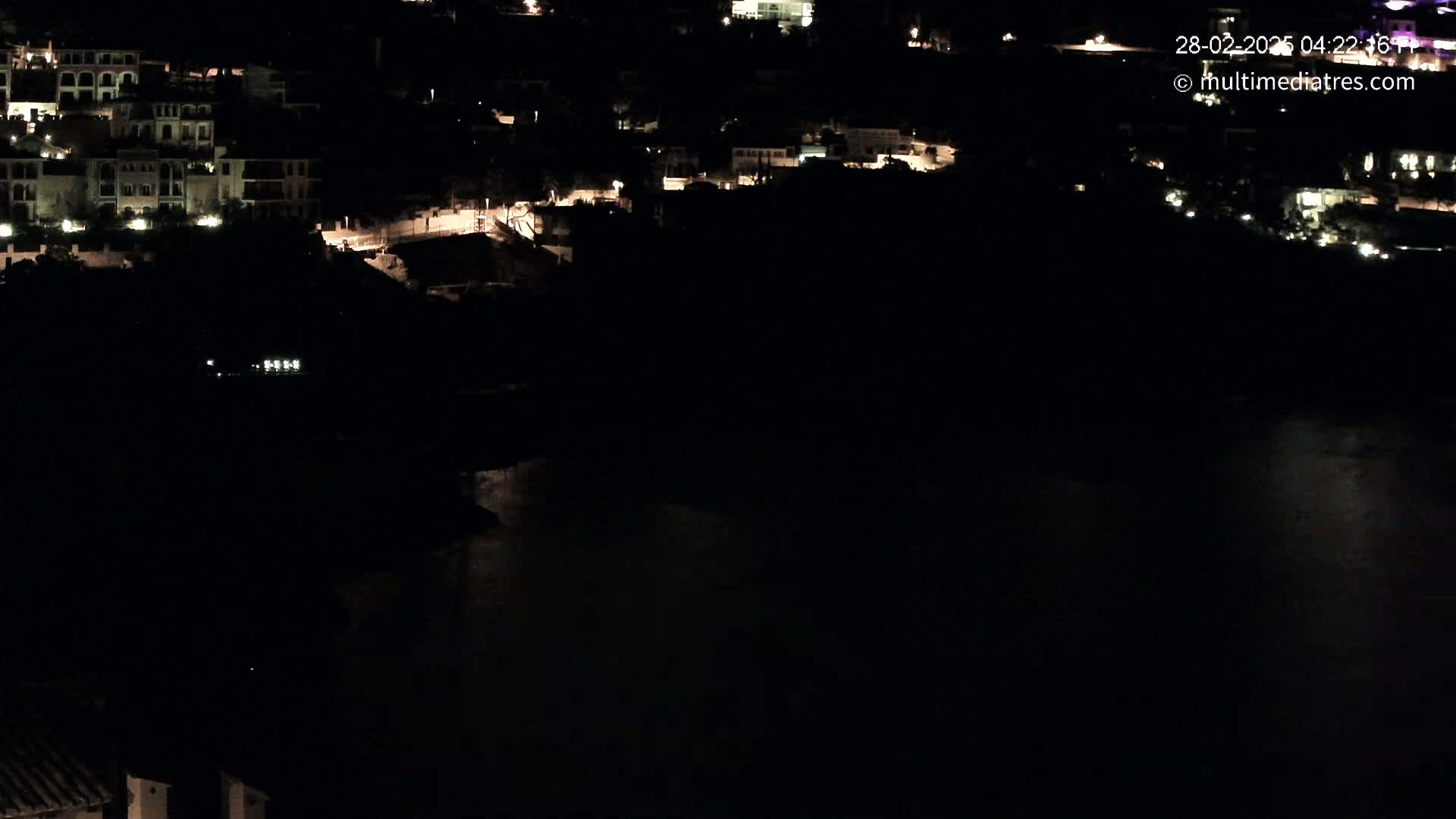 A nighttime view shows a dark body of water below a hill dotted with dimly lit buildings.