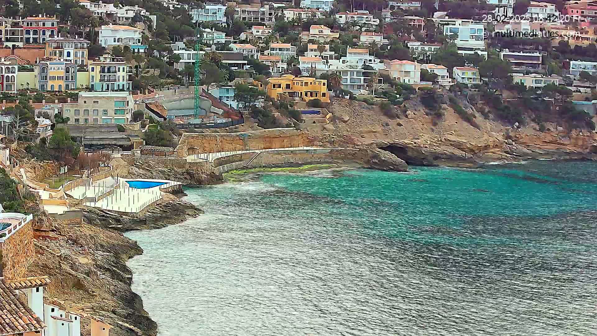 A rocky coastline with turquoise water features numerous houses built into a hillside, a swimming pool, and a partially constructed building.