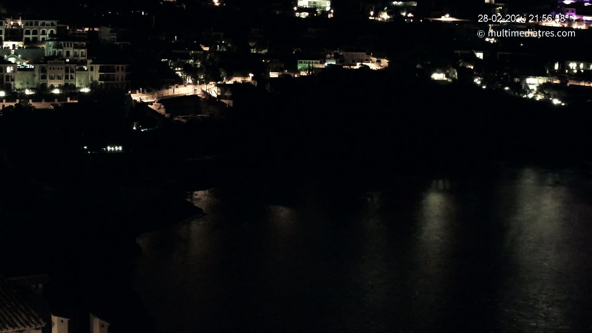 A nighttime, high-angle view of a coastal town with buildings illuminated against a dark body of water.