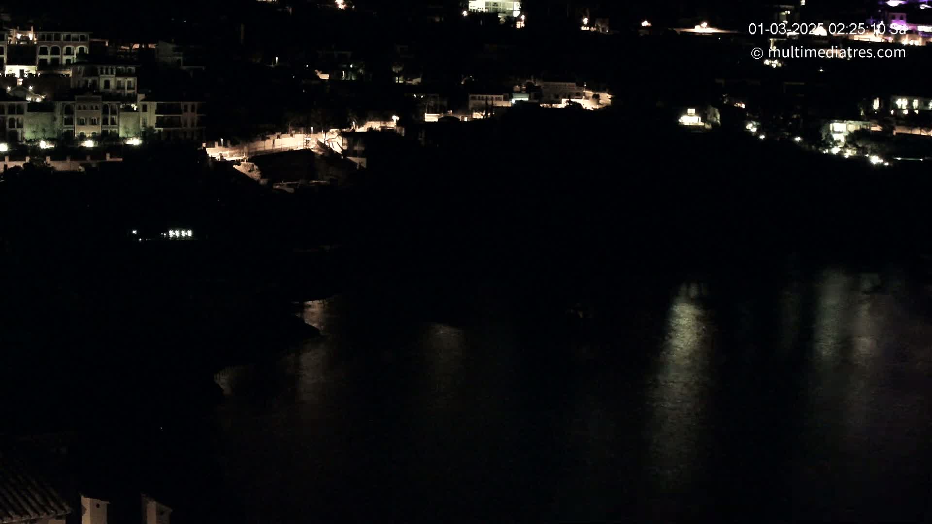A nighttime, high-angle view shows a coastal town with lights illuminating buildings and reflecting on dark water.