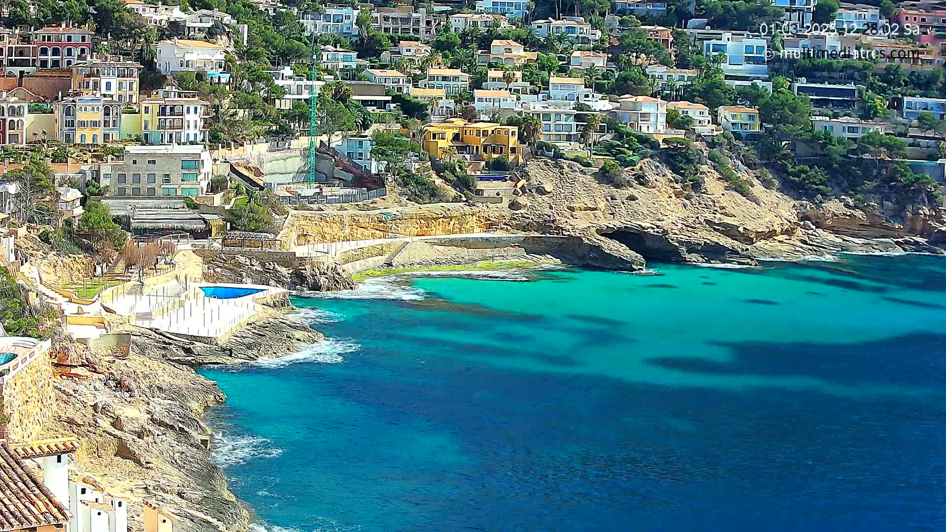 A coastal town with numerous houses clings to a rocky hillside overlooking a vibrant turquoise sea on a sunny day.