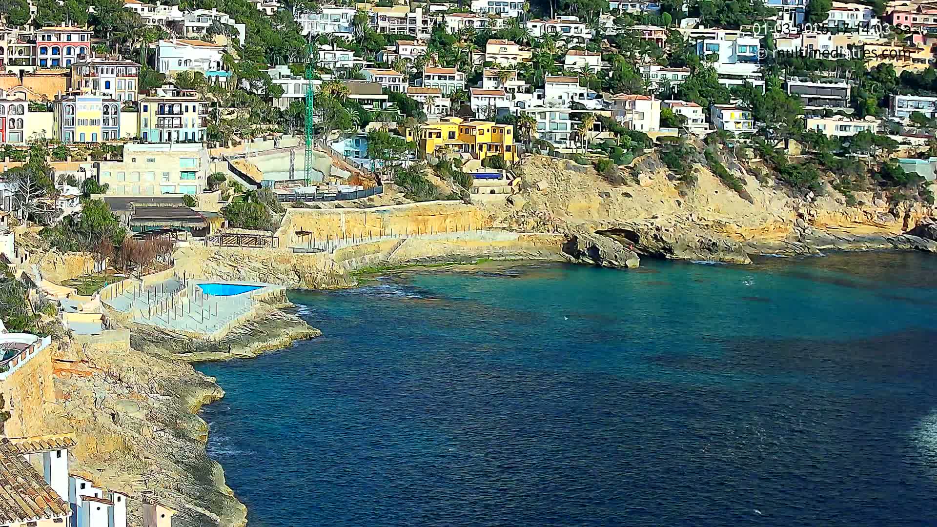 A hillside of Mediterranean-style houses overlooks a rocky coastline and clear turquoise water on a sunny day.