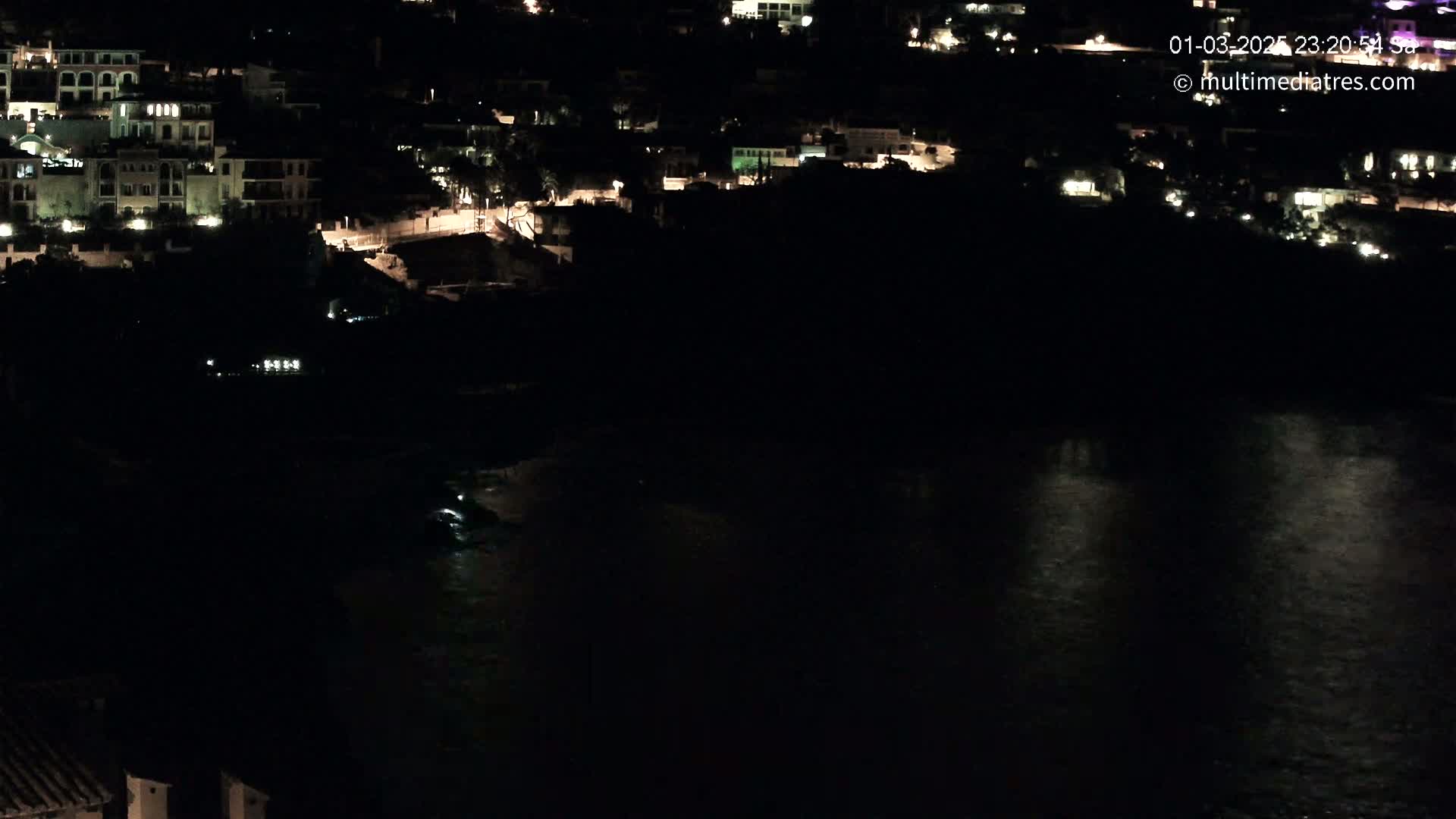 A nighttime view of a coastal town with buildings lit up along a dark body of water.