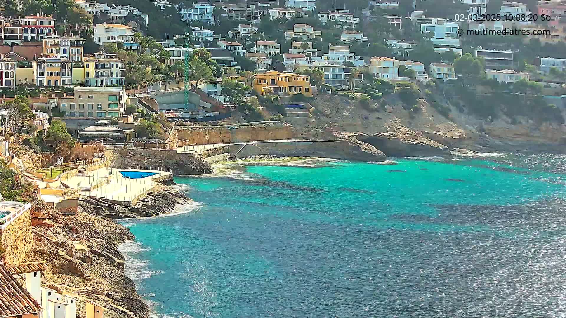 A sunny day reveals a rocky coastline with turquoise water, featuring a small pool and numerous houses built into a hillside.