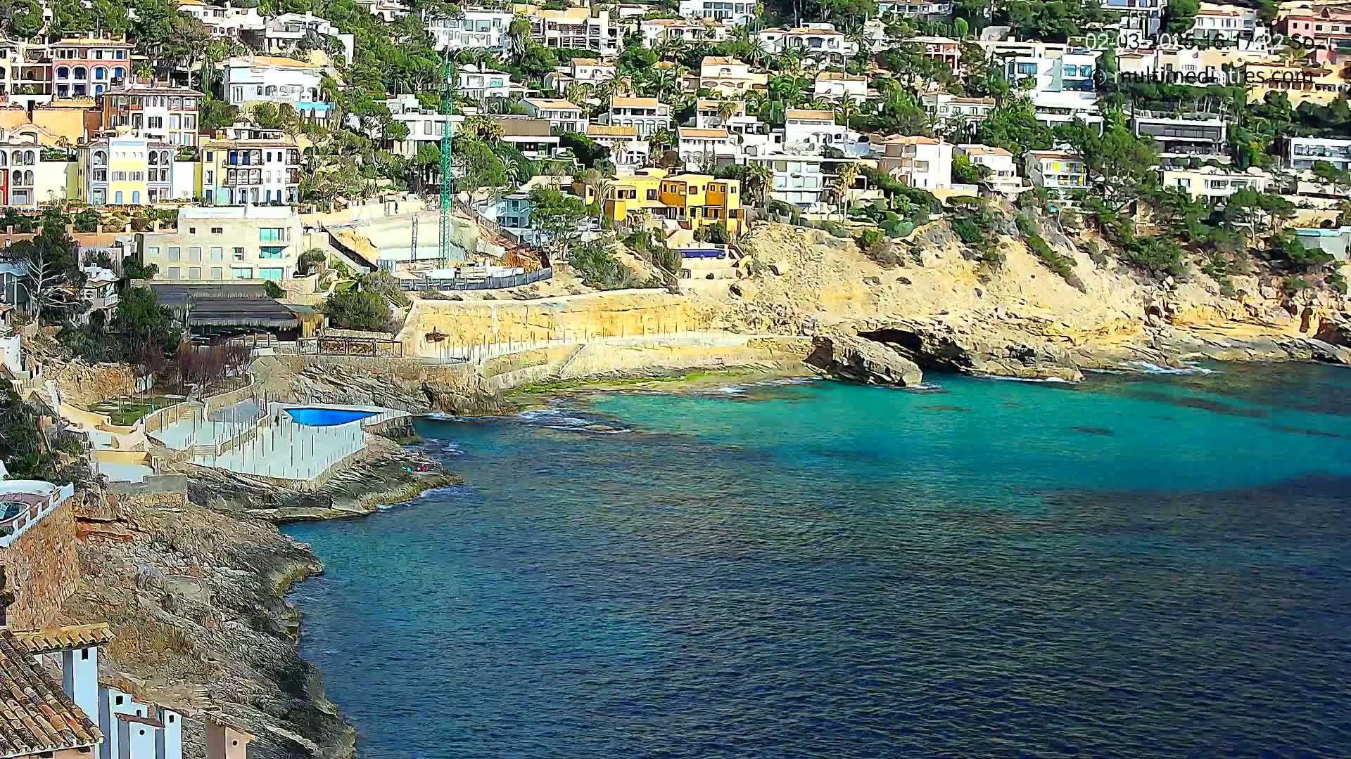 A coastal town with many houses built into a hillside overlooks a calm, turquoise body of water.
