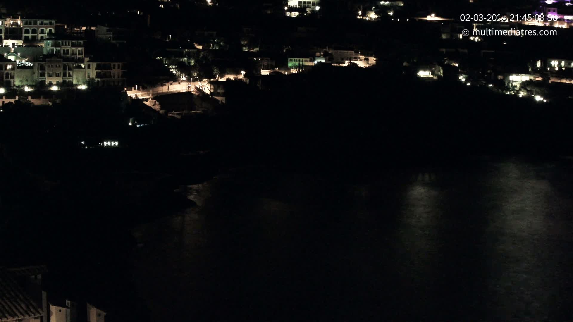 A nighttime, aerial view shows a coastal town with dimly lit buildings and a dark body of water reflecting scattered lights.