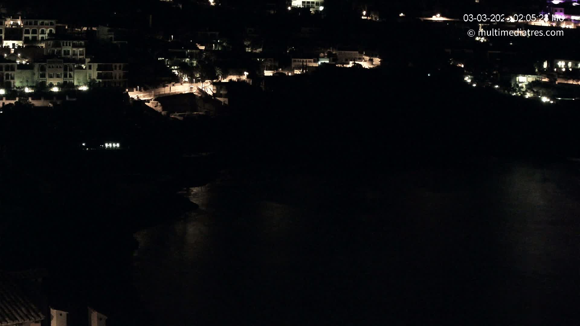A nighttime, aerial view shows a coastal town with dimly lit buildings near a dark body of water.