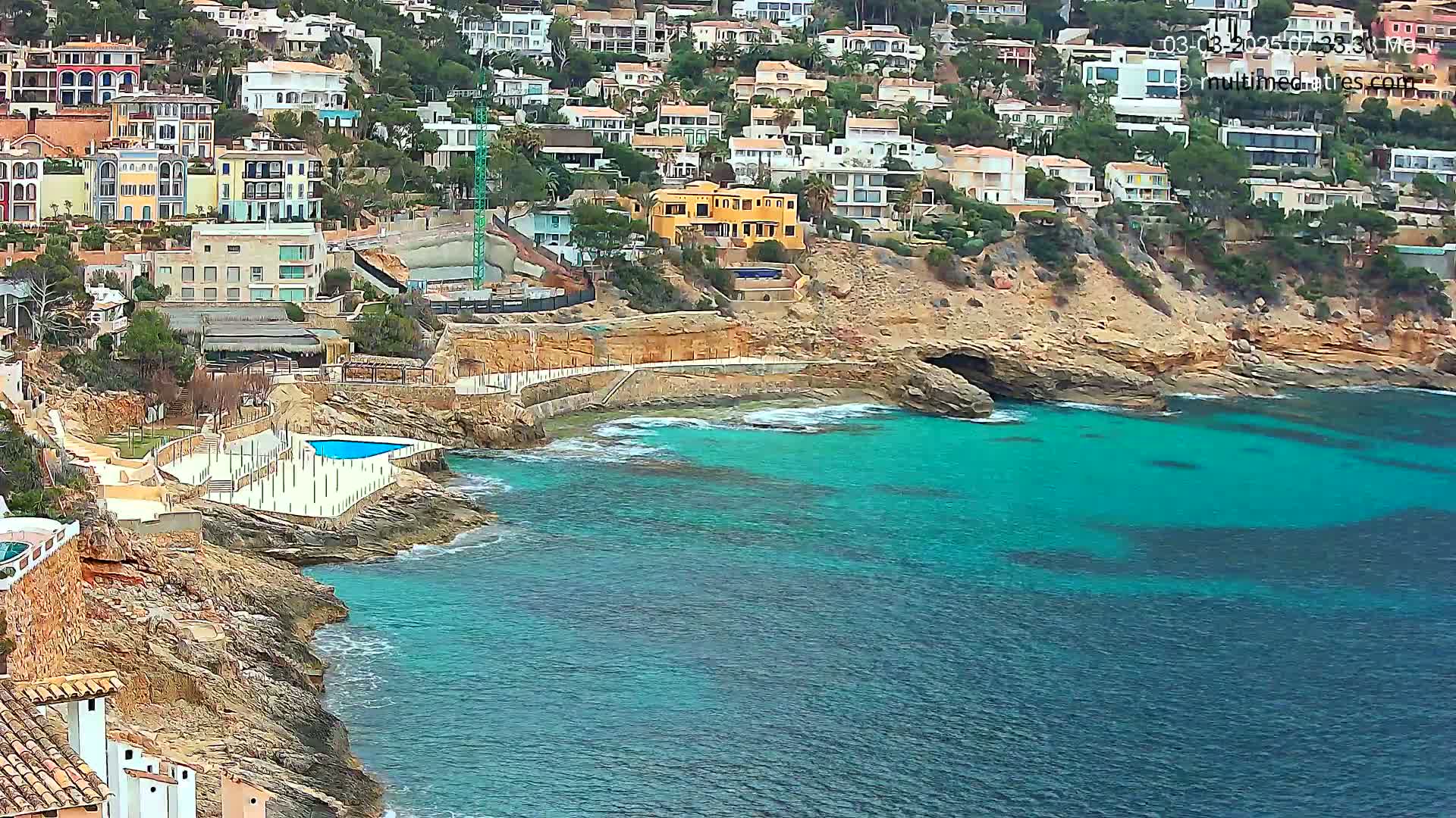 A rocky coastline with turquoise water features a cluster of houses on a hillside, a small swimming pool, and some construction.