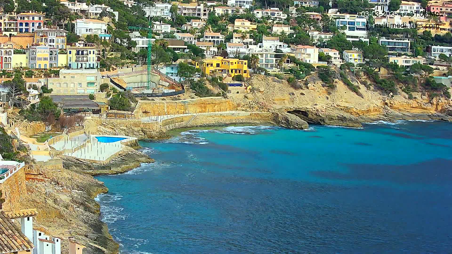 A coastal town with colorful houses built into a hillside overlooking a calm, turquoise ocean with a small swimming pool adjacent to the water.
