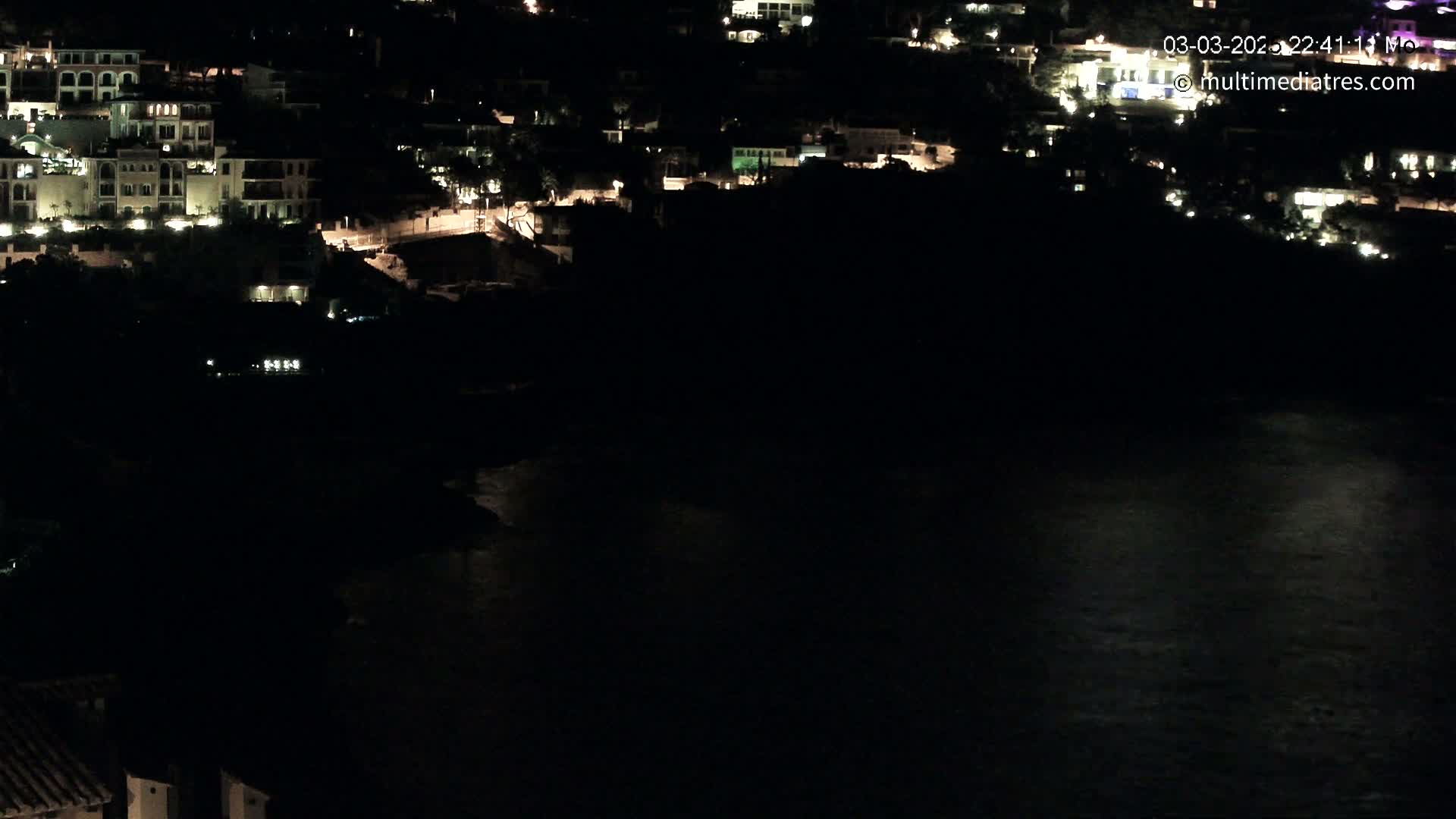 A nighttime, aerial view shows a collection of illuminated buildings situated on a dark hillside overlooking a calm body of water.