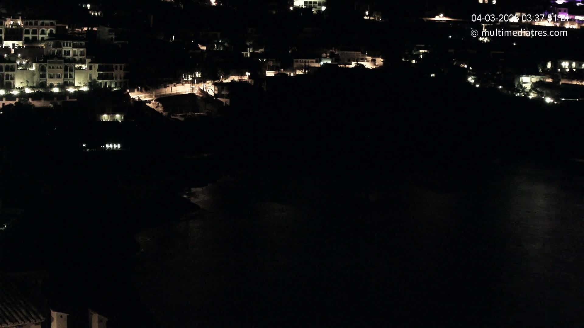 A nighttime view of a coastal town with buildings lit up along the shoreline and dark water below.