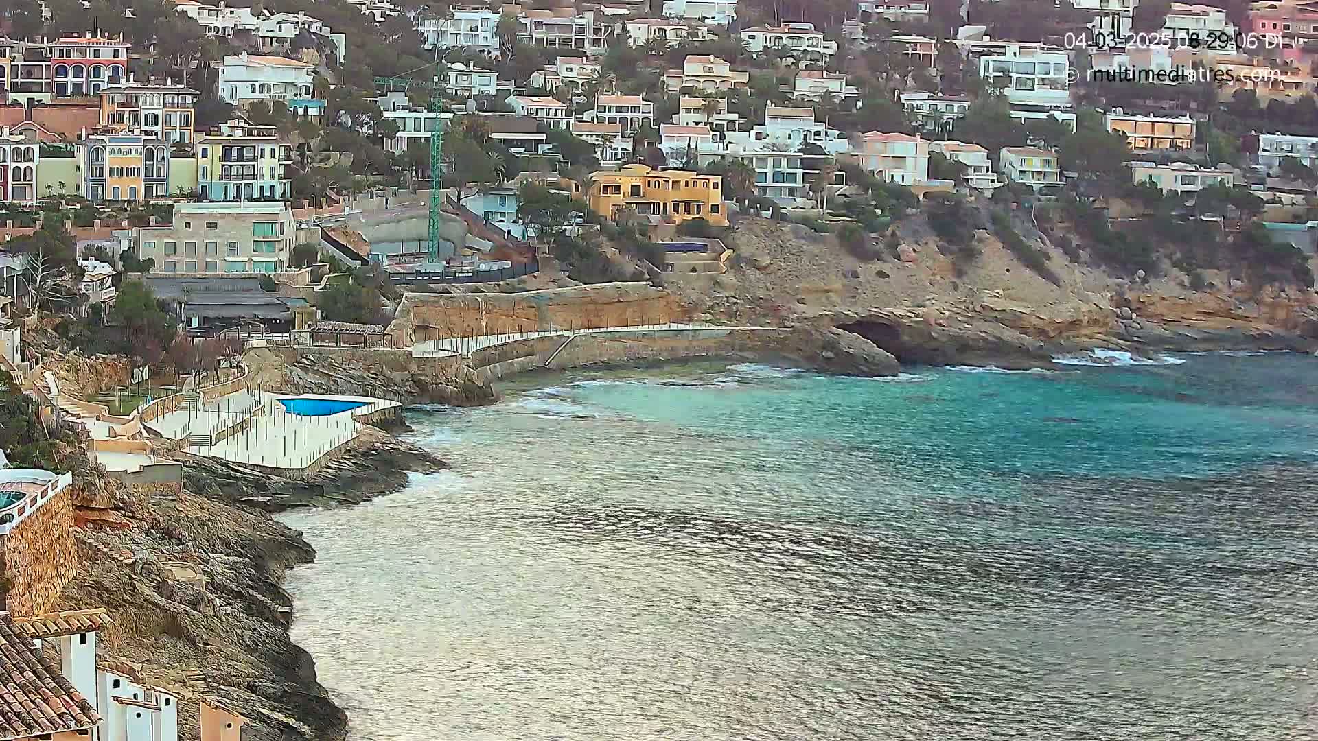A rocky coast with a small cove and a swimming pool overlooks a calm, teal sea, with a hillside densely packed with buildings behind it.