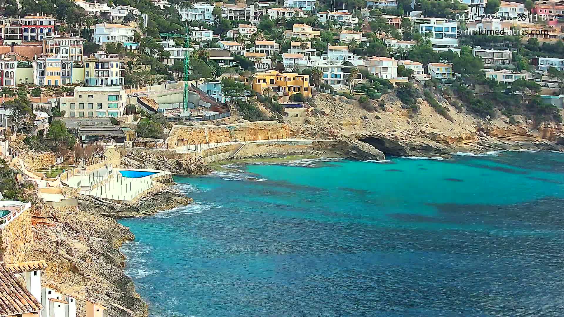 A rocky coastline with turquoise water features numerous houses on a hillside, and a small swimming pool is visible near the water's edge.