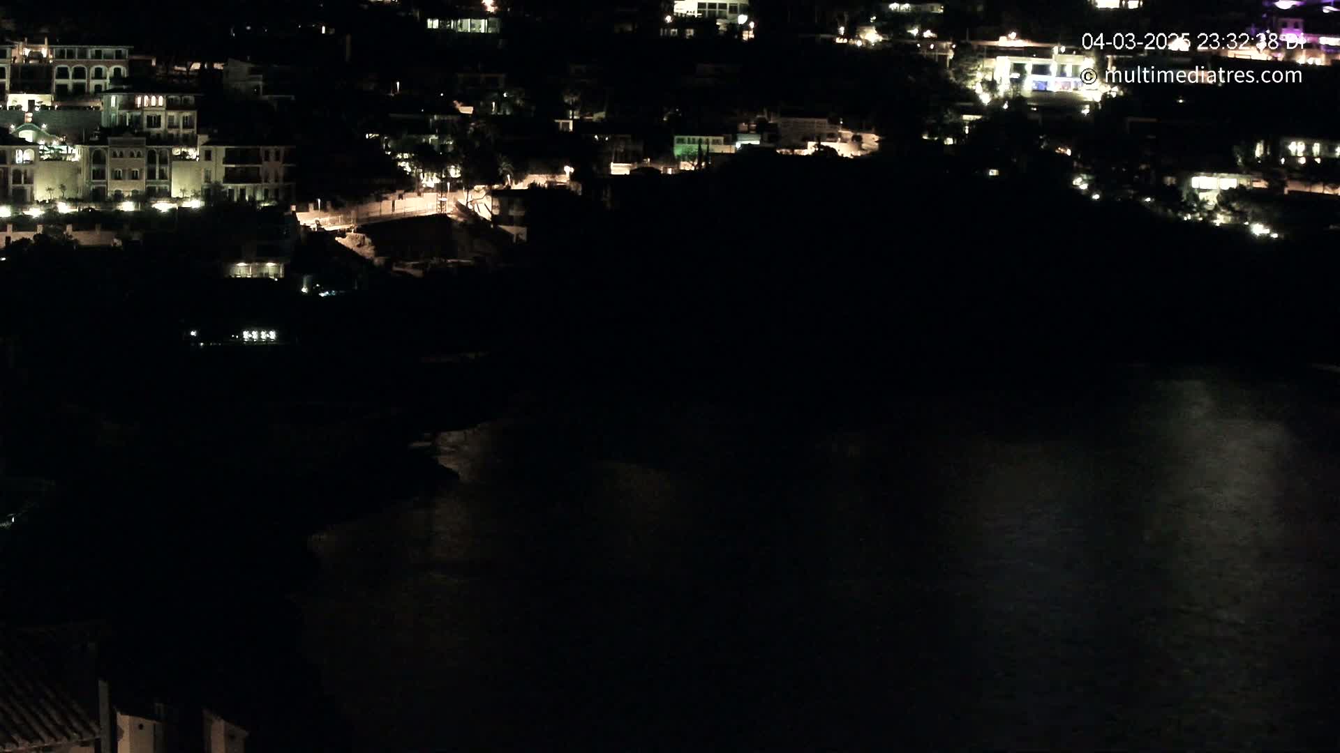 A nighttime aerial view shows a coastal town with dimly lit buildings and a dark body of water.
