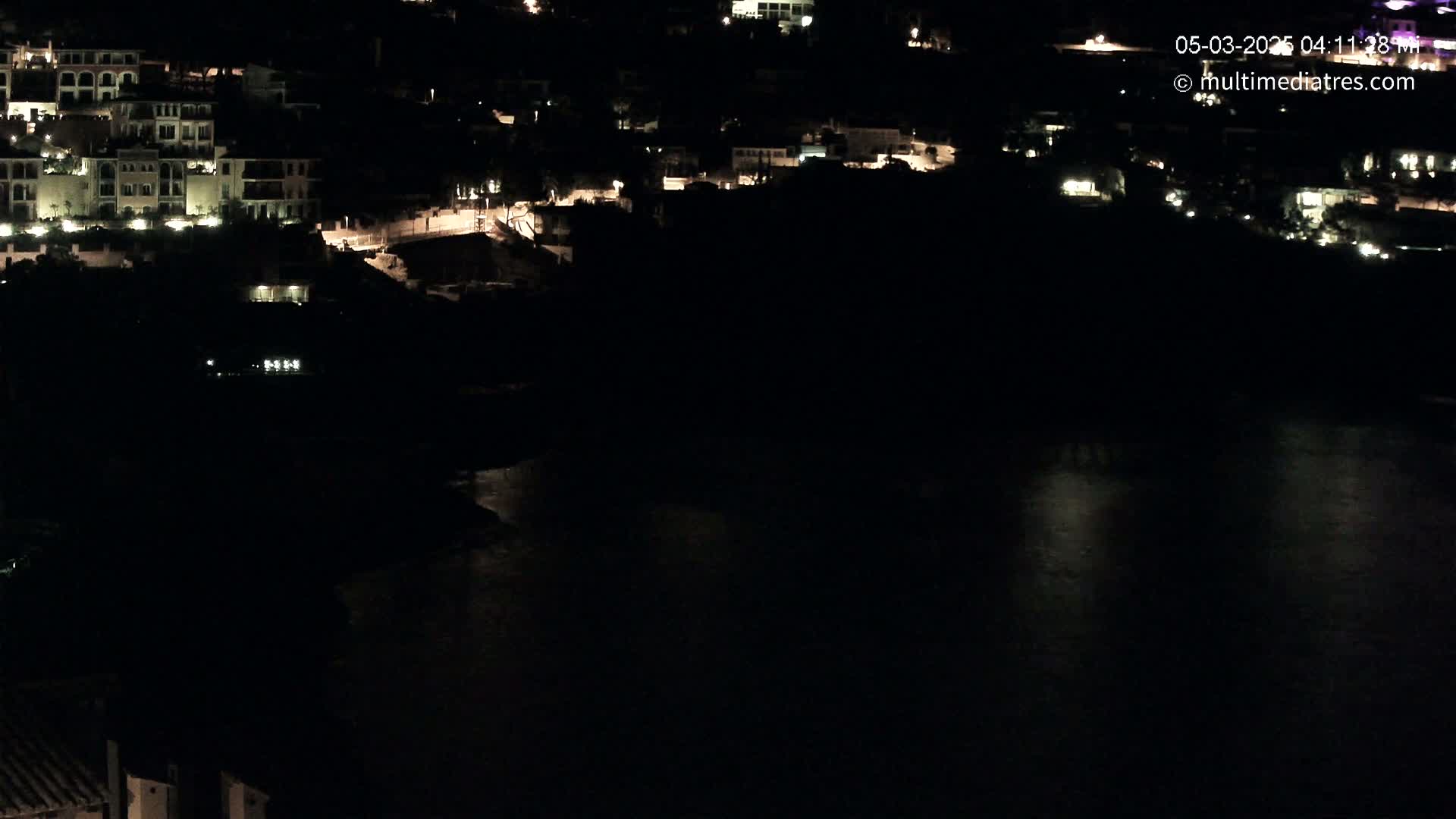 At night, a collection of buildings with illuminated windows sits on a dark hillside overlooking a calm body of water.