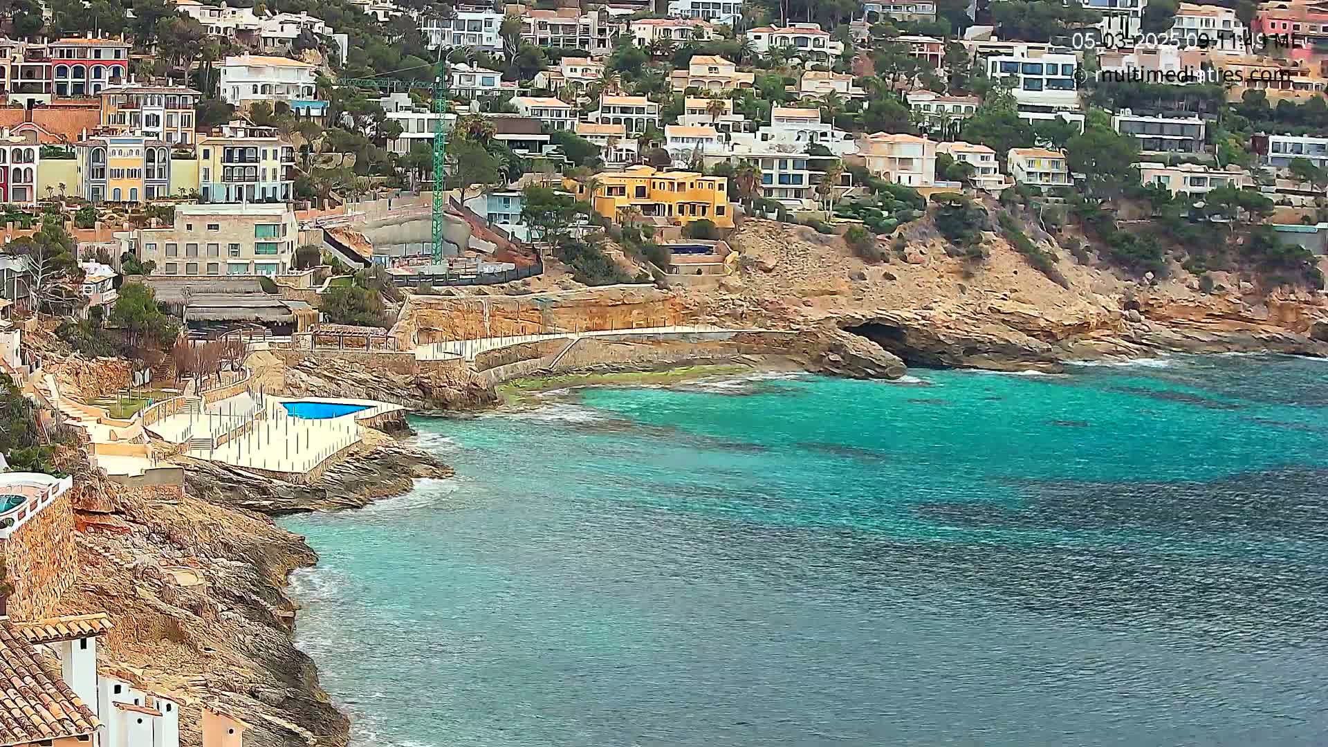 A rocky coastline with turquoise water features a small swimming pool and numerous houses built into a hillside.