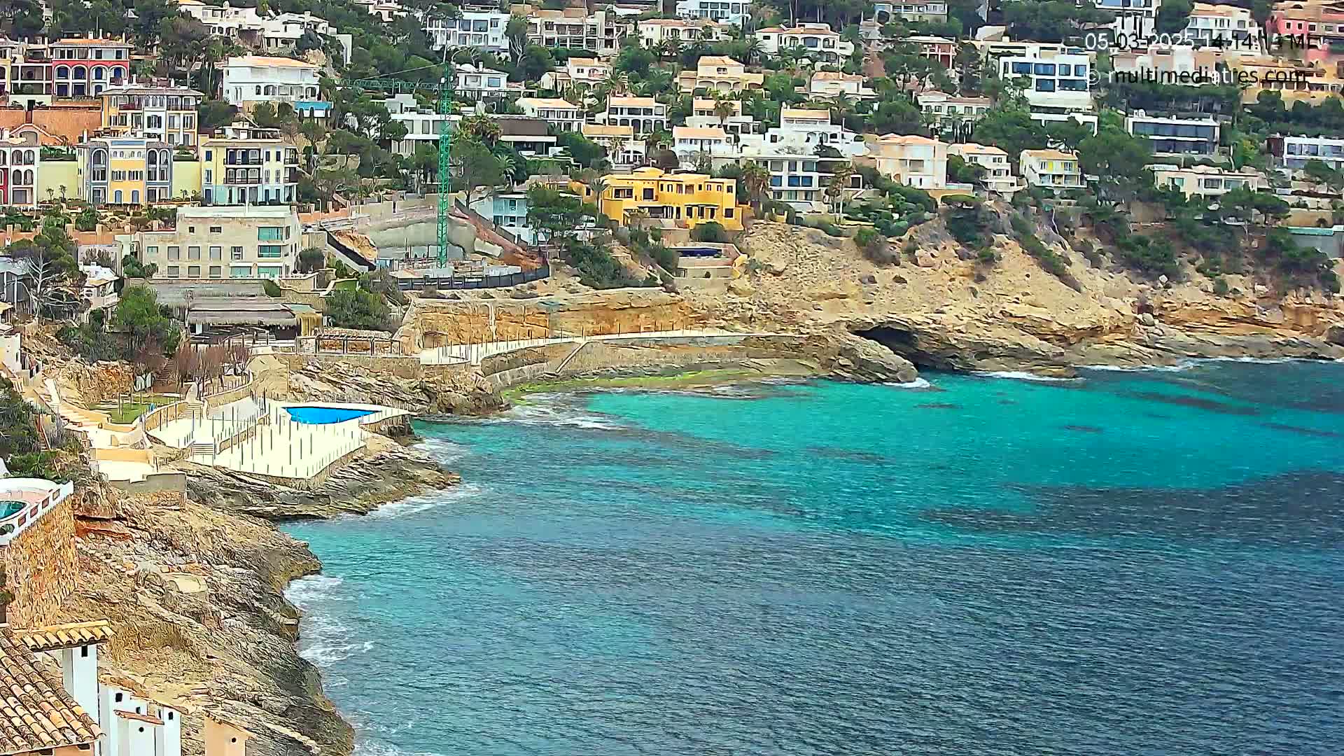 A coastal community of colorful houses on a hillside overlooks a tranquil turquoise bay with a small pool on the rocky shore.