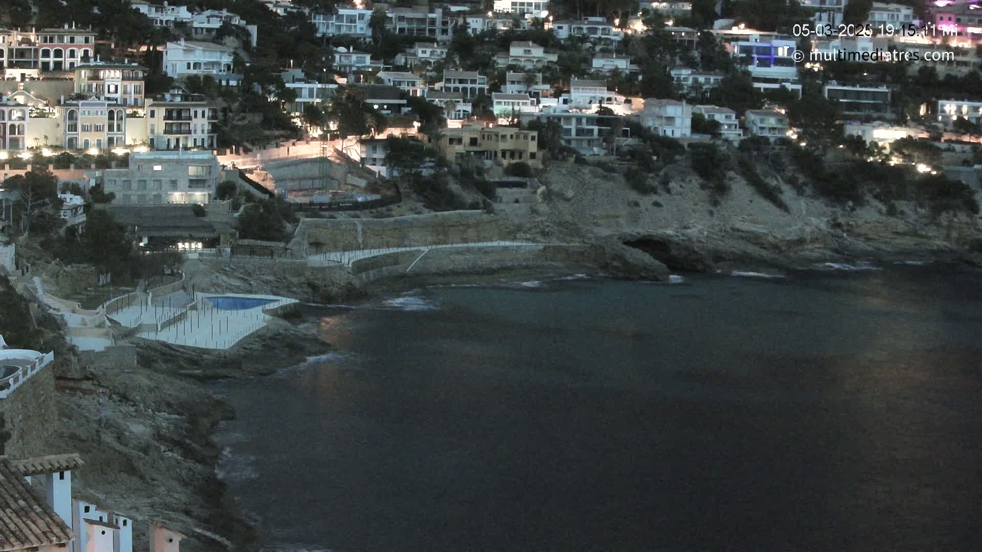 A coastal town at dusk, with houses lit up on a hillside overlooking a dark, calm sea.