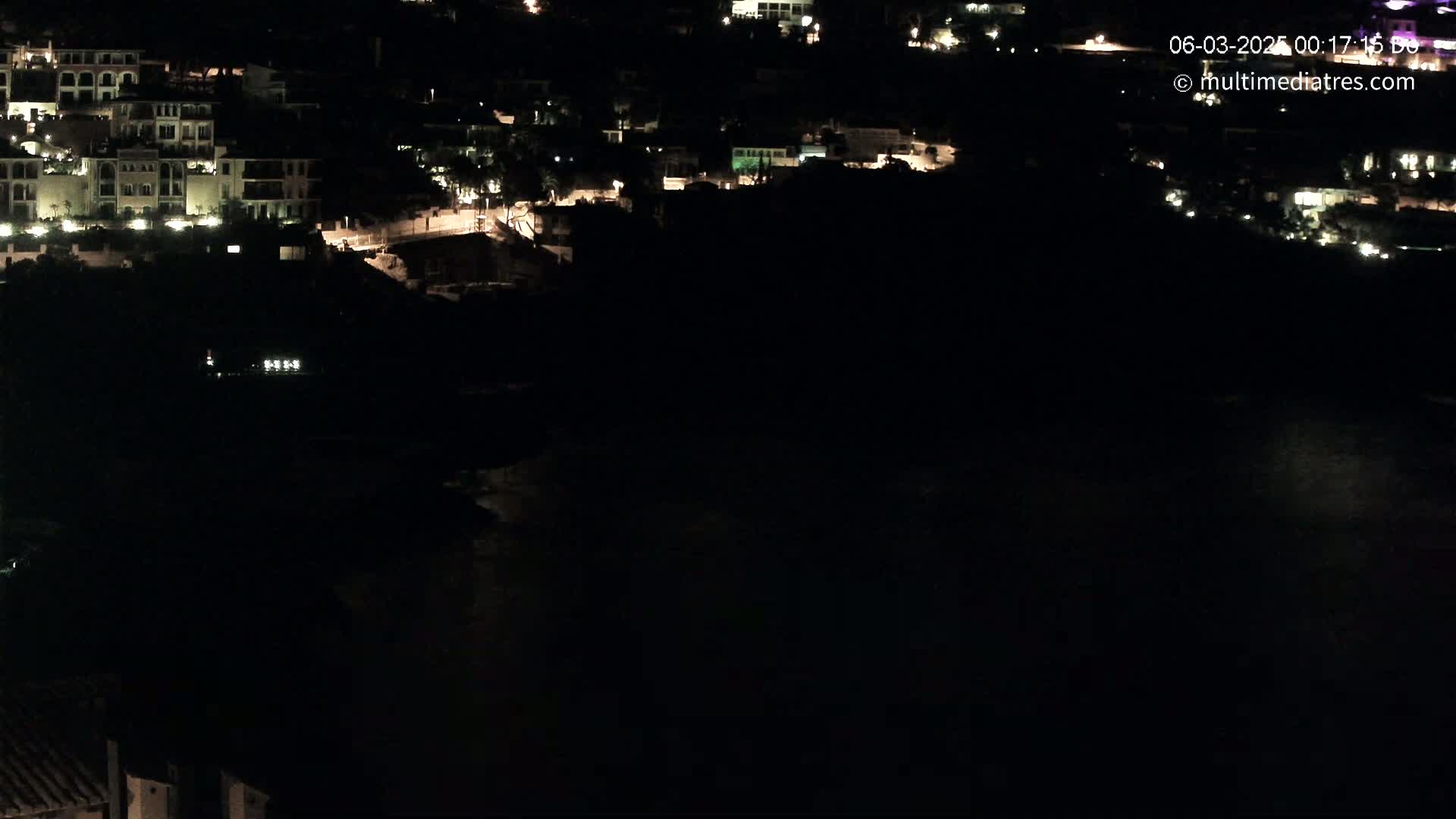 A nighttime, high-angle view shows a collection of illuminated buildings nestled in dark, hilly terrain near a body of dark water.