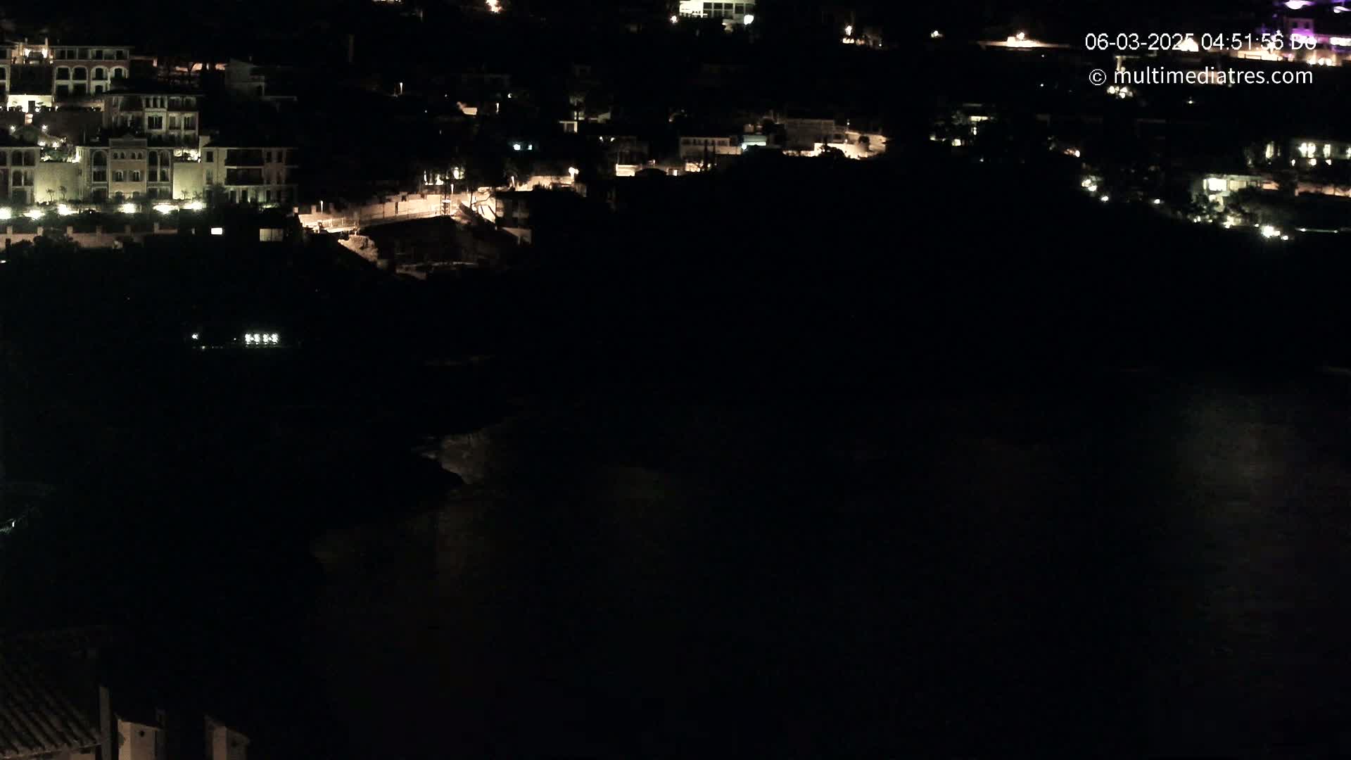 A nighttime, aerial view shows a coastal town with dimly lit buildings and a dark body of water.