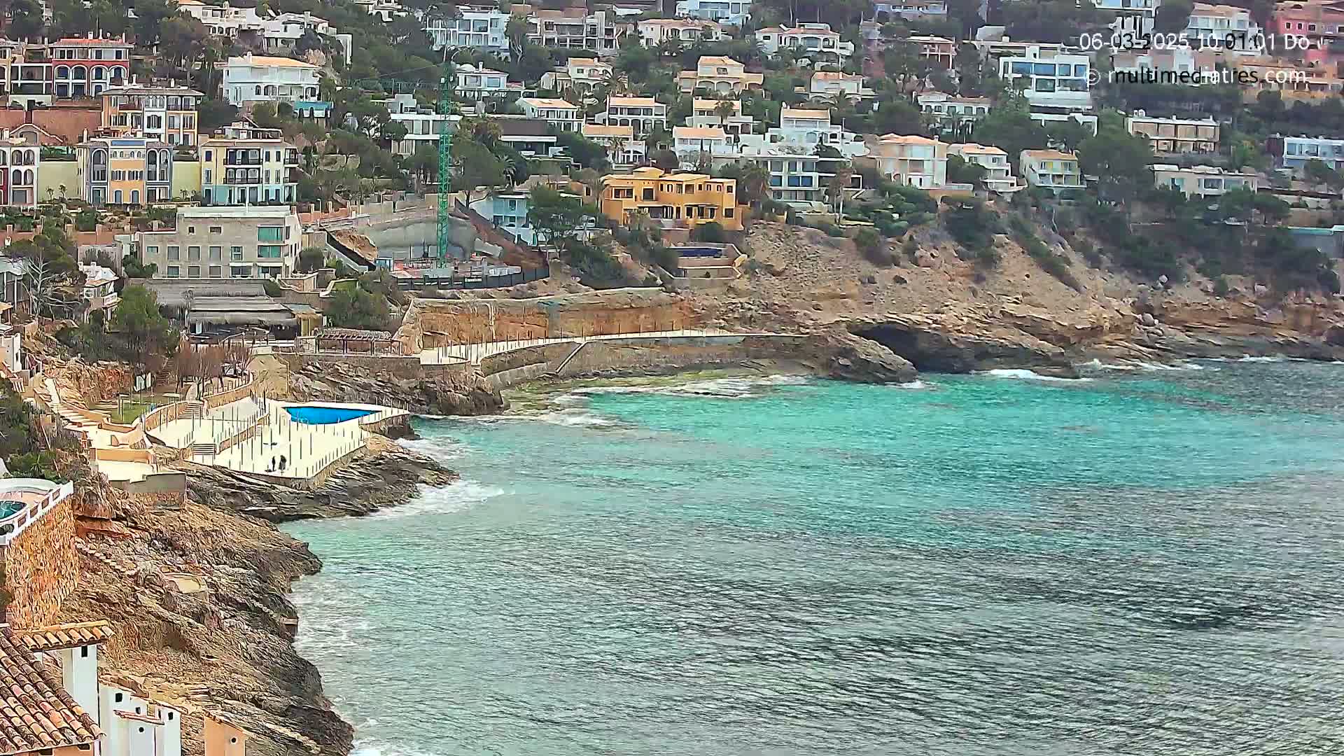 A rocky coastline with turquoise water features a cluster of houses on a hillside, a small swimming pool, and a paved walkway.