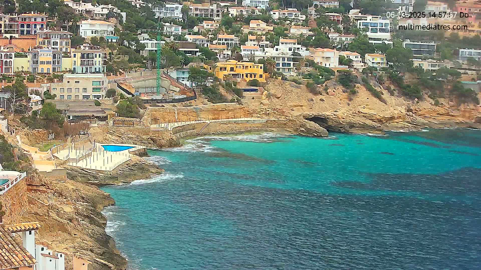 A rocky coastline with teal water features a small swimming pool and many houses built into the hillside.