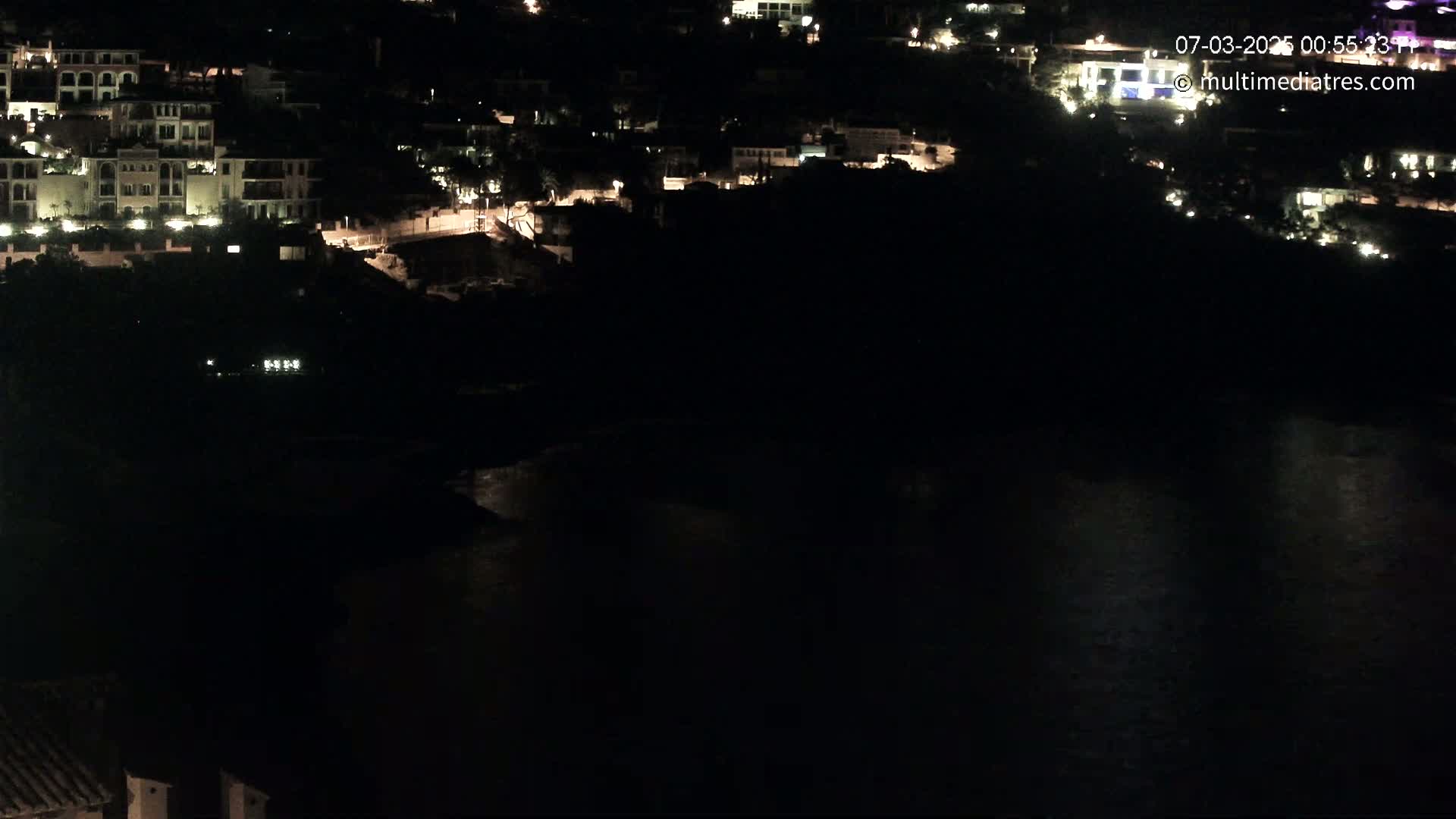 A nighttime view of a coastal town with illuminated buildings and a dark body of water below.