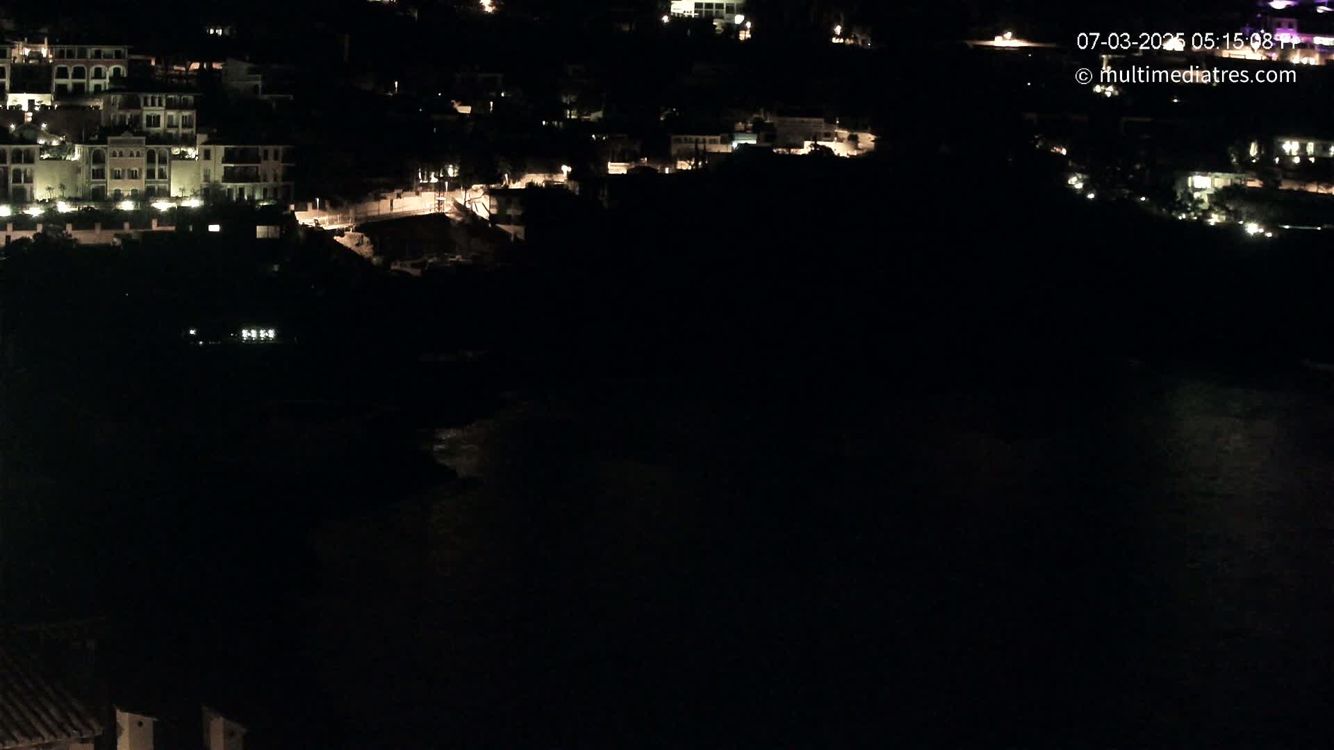 A nighttime, high-angle view shows a dark body of water next to a hillside of dimly lit buildings.