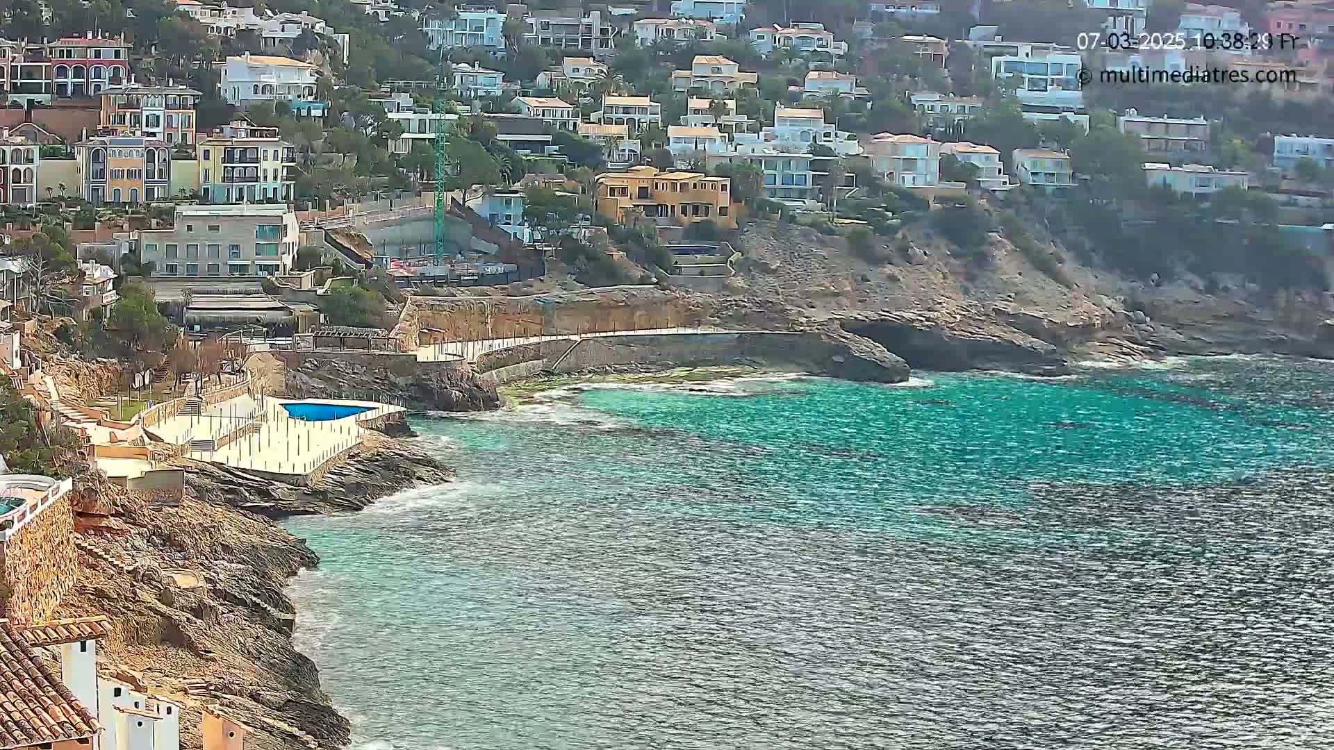 A rocky coastline with turquoise water features a small pool and numerous houses on a hillside under sunny conditions.