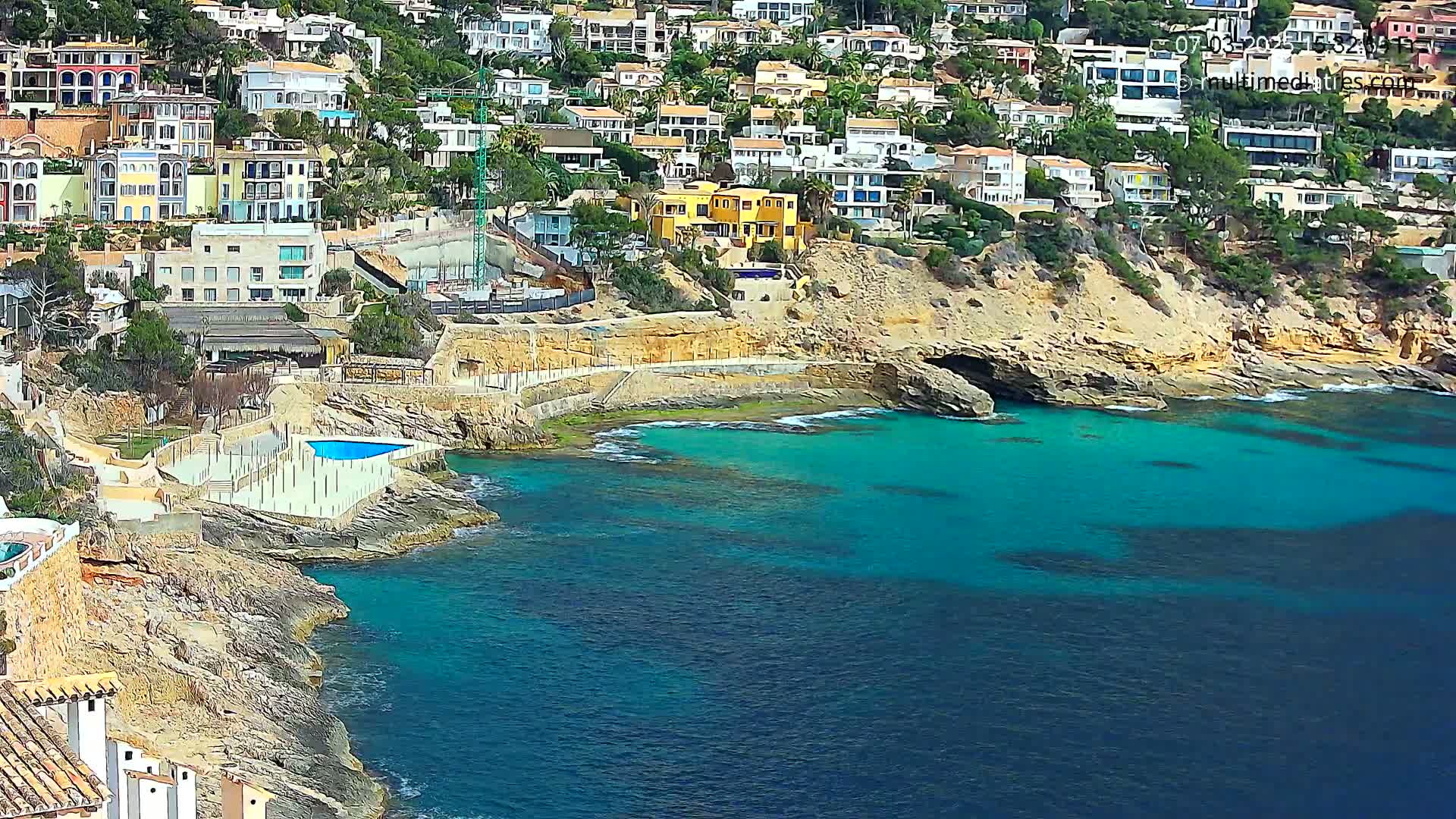 A rocky coastline with numerous houses cascading down a hillside meets a vibrant teal sea on a sunny day.