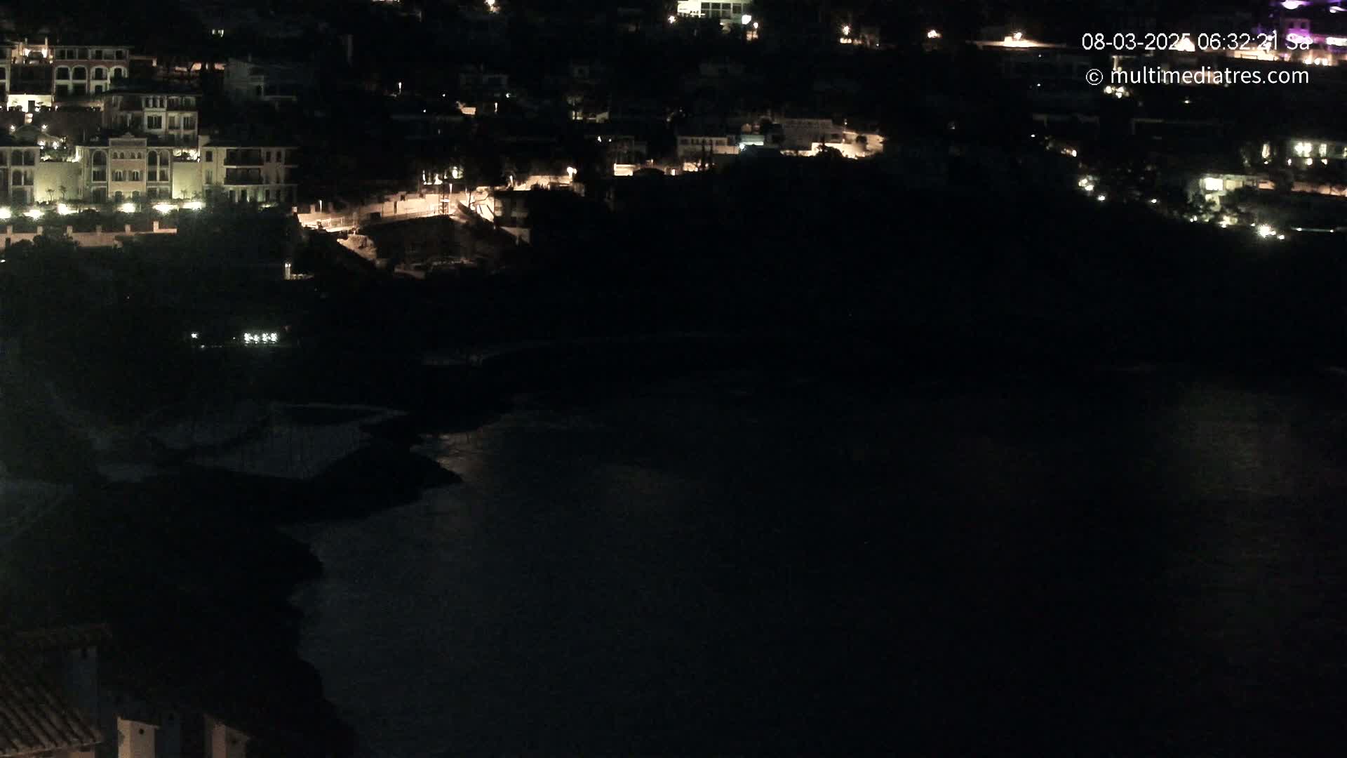 A nighttime view shows a dark body of water below a hillside of buildings with some lights on.