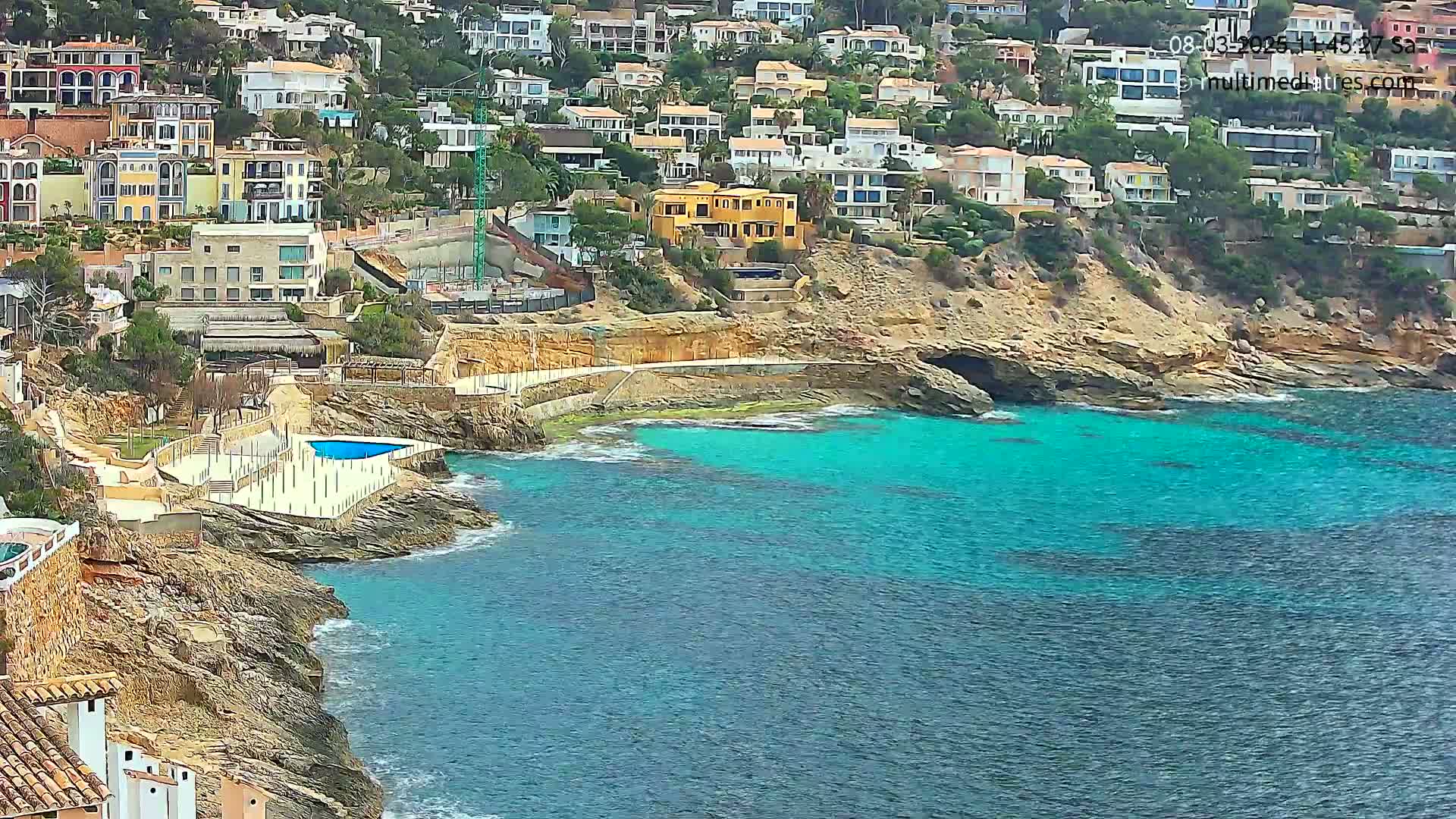 A rocky coastline with turquoise water features a small swimming pool and many houses built into the hillside.