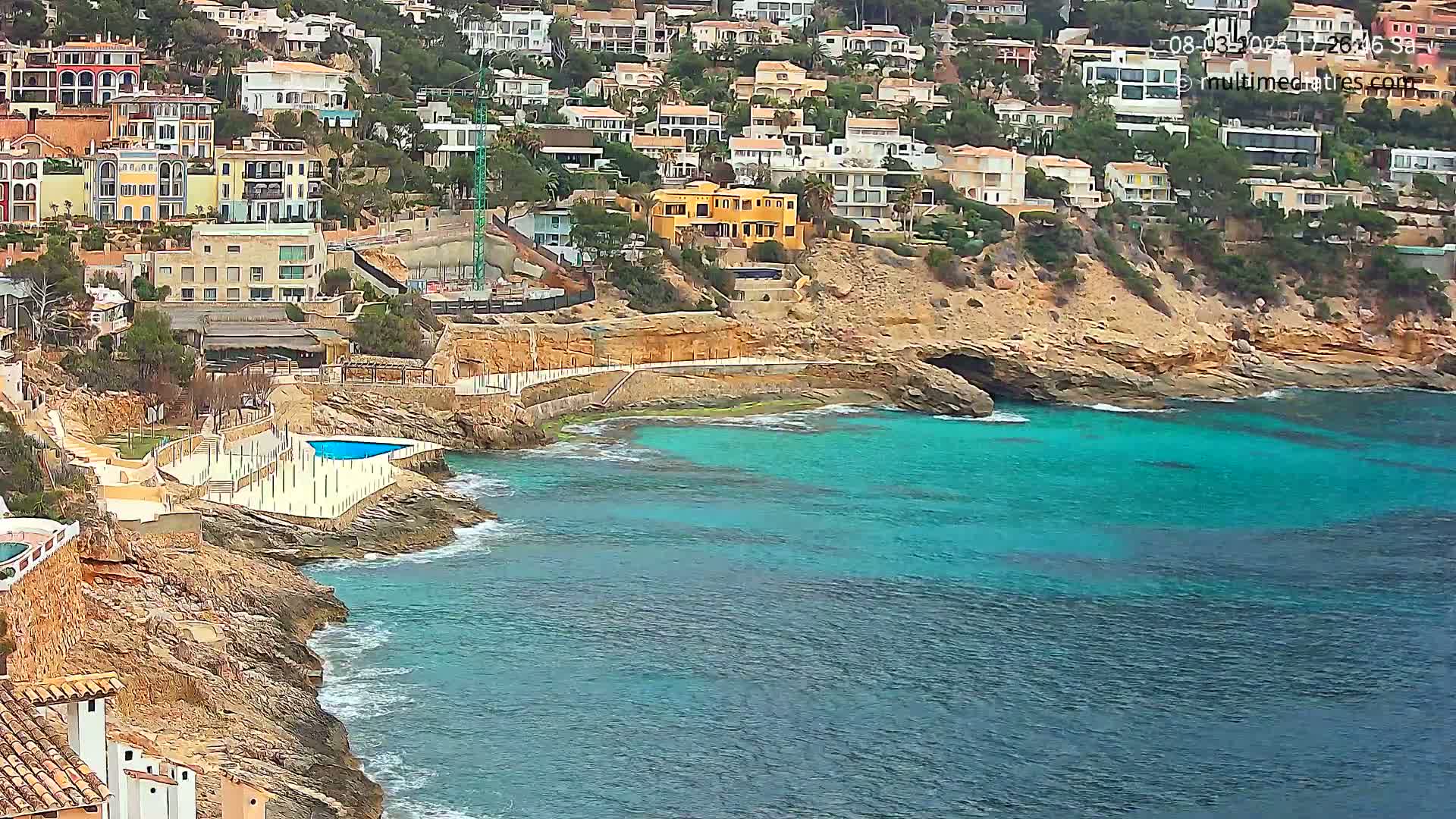 A rocky coastline with turquoise water features numerous houses on a hillside, a small pool, and some light wave action.