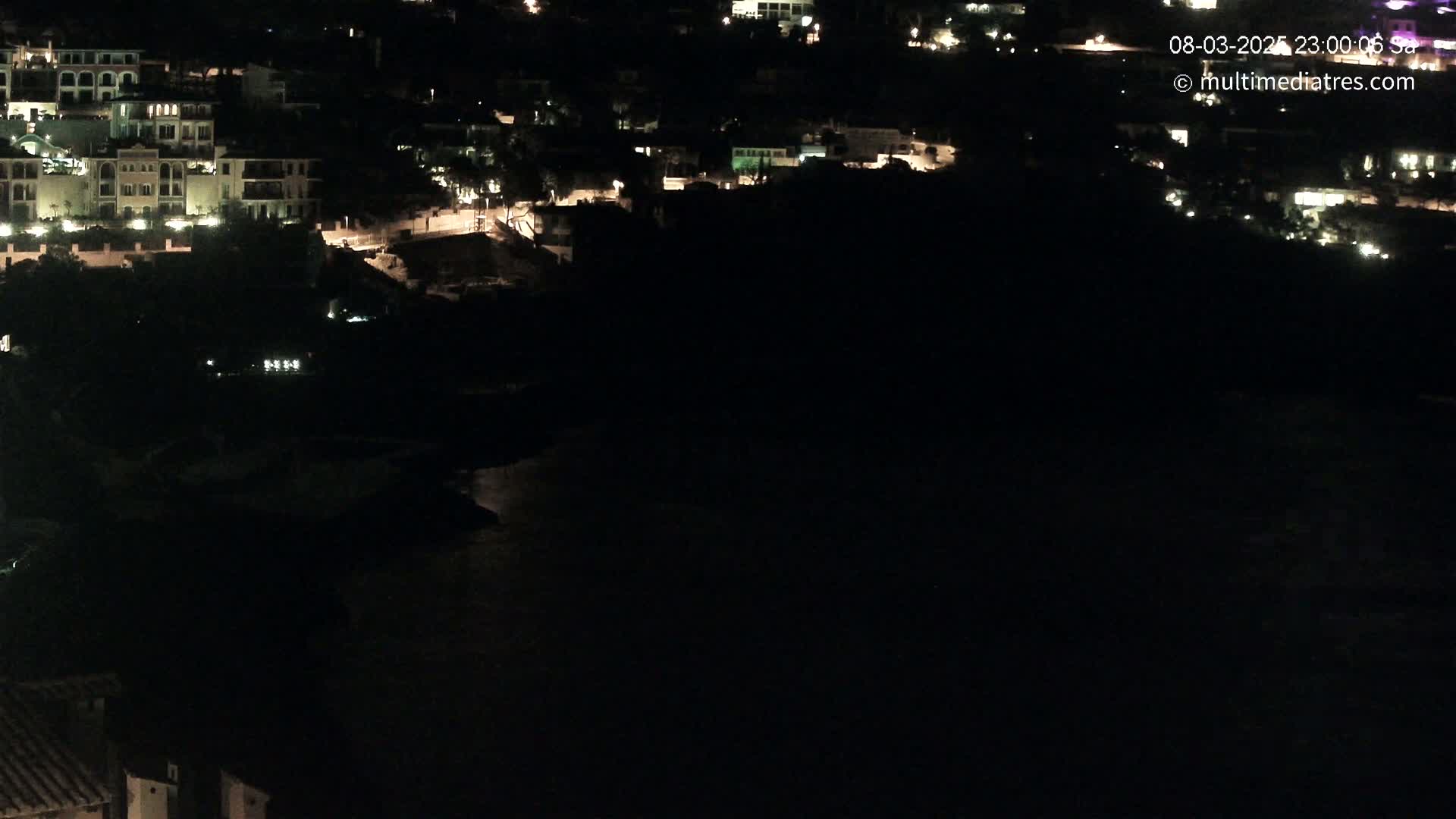 A nighttime, aerial view shows a coastal town with illuminated buildings next to a dark body of water.