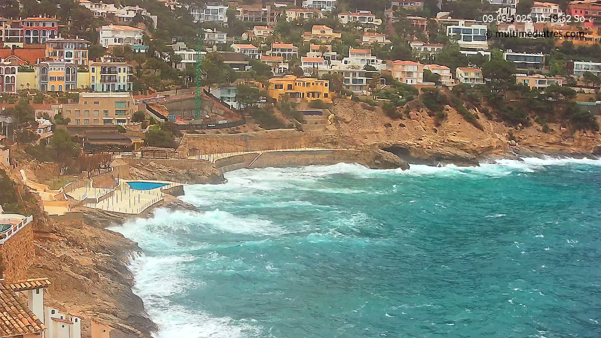 A rocky coastline with a cluster of buildings is battered by rough, turquoise waves under an overcast sky.